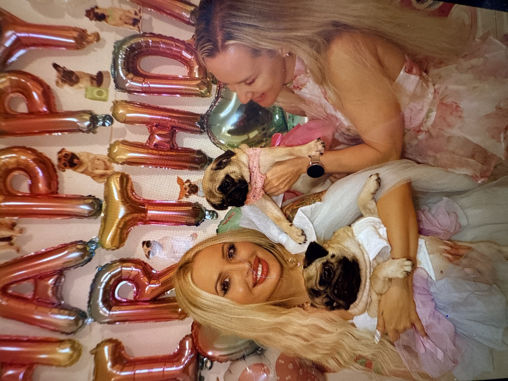 This is a vibrant, brightly lit indoor portrait featuring two smiling blonde women and two pugs, set against a backdrop of festive decorations. The image is oriented vertically, with the subjects positioned diagonally.
The woman at the top of the frame, with long, light blonde hair pulled back at the crown, is shown from the chest up, looking down with a warm smile at a pug she cradles. She wears a light-colored, possibly floral-patterned dress. The pug she holds is dressed in a pink knitted top and appears calm. A smartwatch is visible on her left wrist.
The second woman, positioned in the lower part of the frame, has long, wavy blonde hair and a radiant smile, looking directly towards the viewer. She holds another pug, which is wearing a white top and a pastel-colored, possibly tulle, skirt. This woman is dressed in a white top and a similar ruffled, multi-colored (pink, white, and purple) tulle skirt or dress.
The background is dominated by large, shiny foil balloons in rose gold and gold colors, spelling out "HAPPY BIRTHDAY" vertically. Numerous small pug figurines or cutouts are scattered among the balloons, enhancing the celebratory and pet-themed atmosphere. Both women appear happy and affectionate towards their pugs, indicating a joyful occasion, likely a birthday party. The setting suggests a festive daytime event indoors.