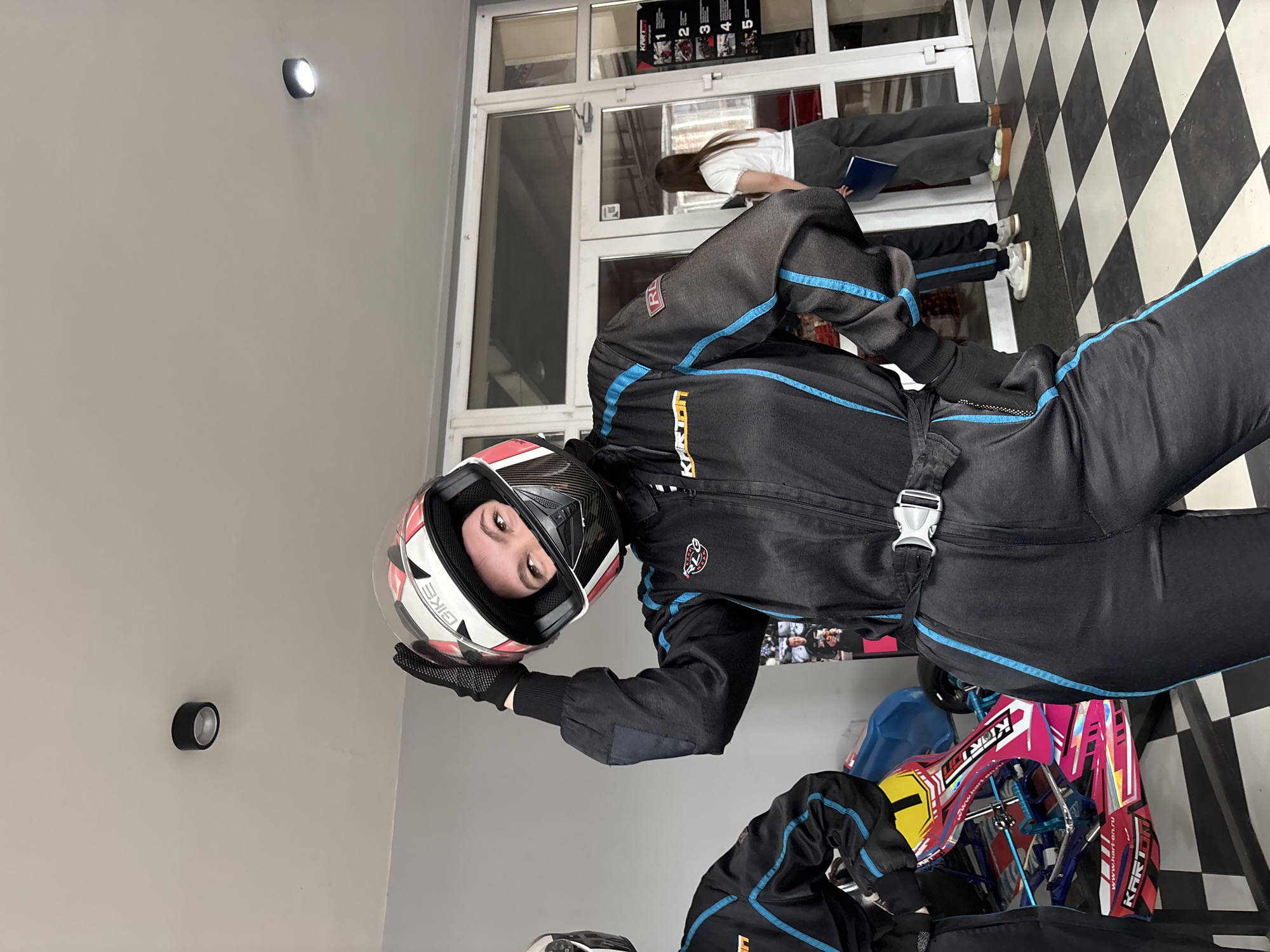 A young person wearing a black racing suit and a helmet is standing indoors. The person is holding the helmet with a gloved hand. In the background, there is a black and white checkered floor, a doorway with people standing near it, and a kart. The photograph was taken in Perm, Russia.