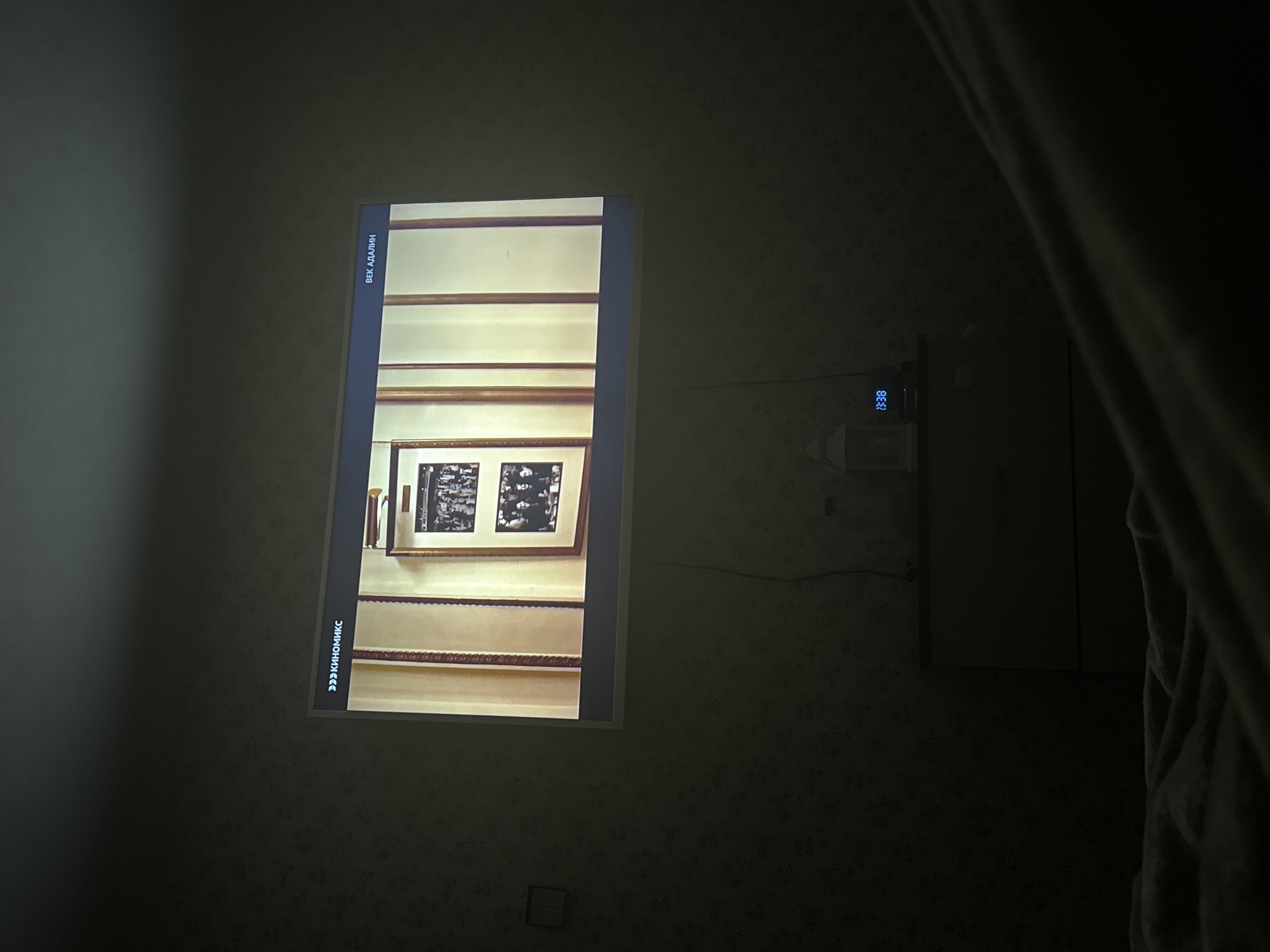 A television screen displays an image of a hallway with framed pictures, while also showing the time as 19:38. The screen is mounted on a patterned wall in a dimly lit room, and a portion of a curtain is visible on the right side of the image. The location is unknown.