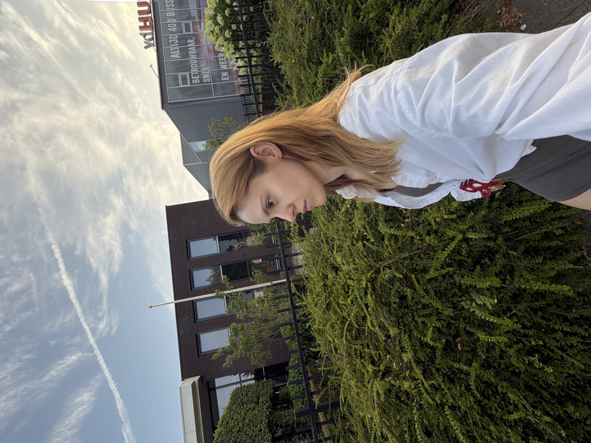 A woman with blonde hair is standing outside in front of a building and lush green bushes. The building has dark brick and large windows. On the building's facade, there is a sign with red lettering that reads "VDHU" and below that, in smaller white text, "ALTIJD 400 BUSSIMMERTJES" and "BETROUWBAAR, SNEL". The woman is wearing a white button-down shirt over a dark top and a dark skirt or shorts. She is looking to her left, with a slight frown on her face. The sky above is blue with wispy white clouds and a faint contrail. The overall lighting suggests it is daytime, possibly late afternoon or early morning. The scene is located in the city of Helmond, Netherlands.