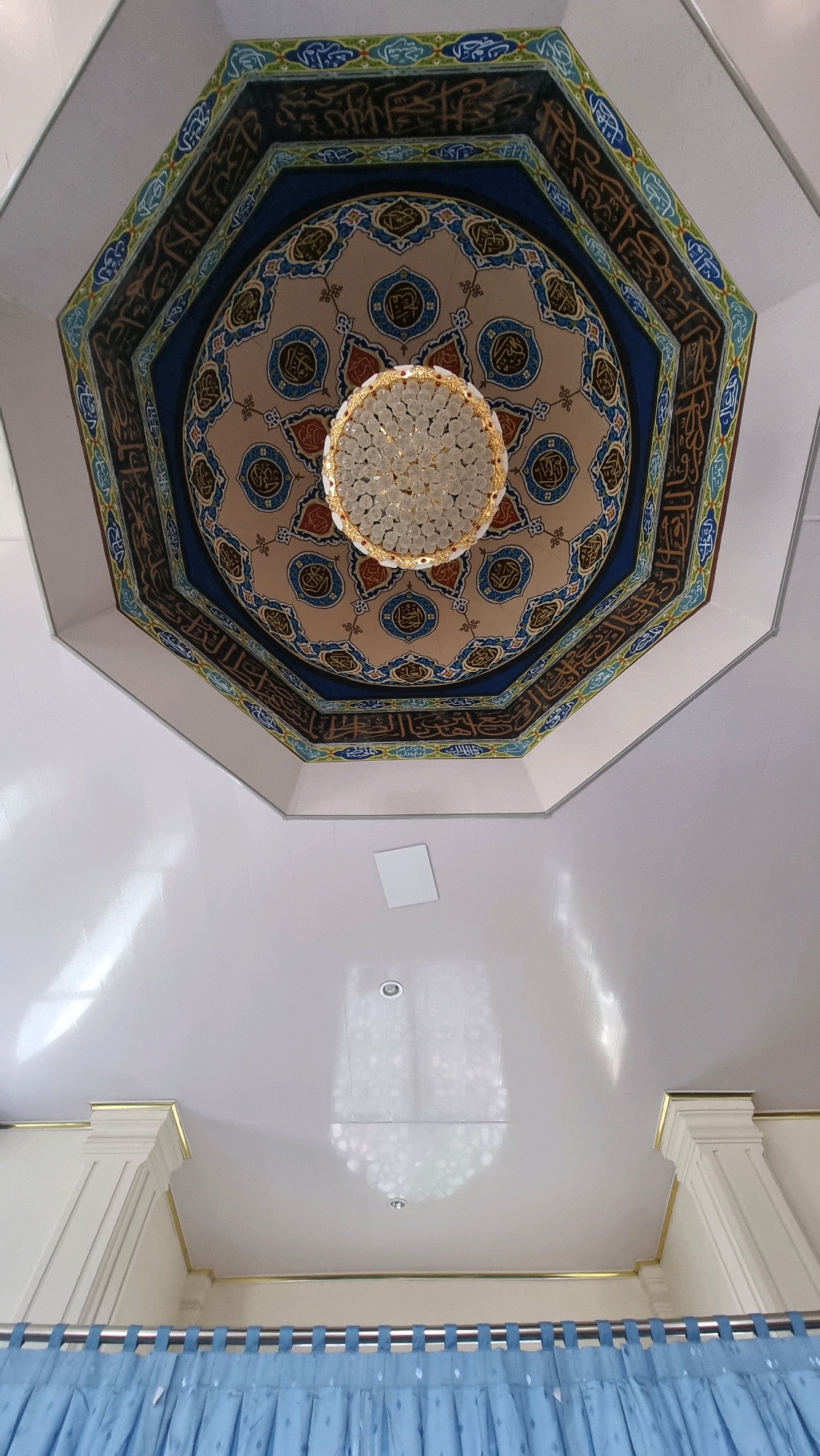 The image shows an interior view looking upwards at an octagonal ceiling with intricate Islamic calligraphy and geometric patterns. In the center of the ceiling is a decorative chandelier. The ceiling extends outwards to a white, flat surface with some light fixtures. The lower portion of the image shows the top of blue curtains hanging from a metal rod, and decorative white pillars with gold trim. The scene appears to be an indoor public space, likely a mosque or a similarly decorated building in Lhoksukon, Indonesia.