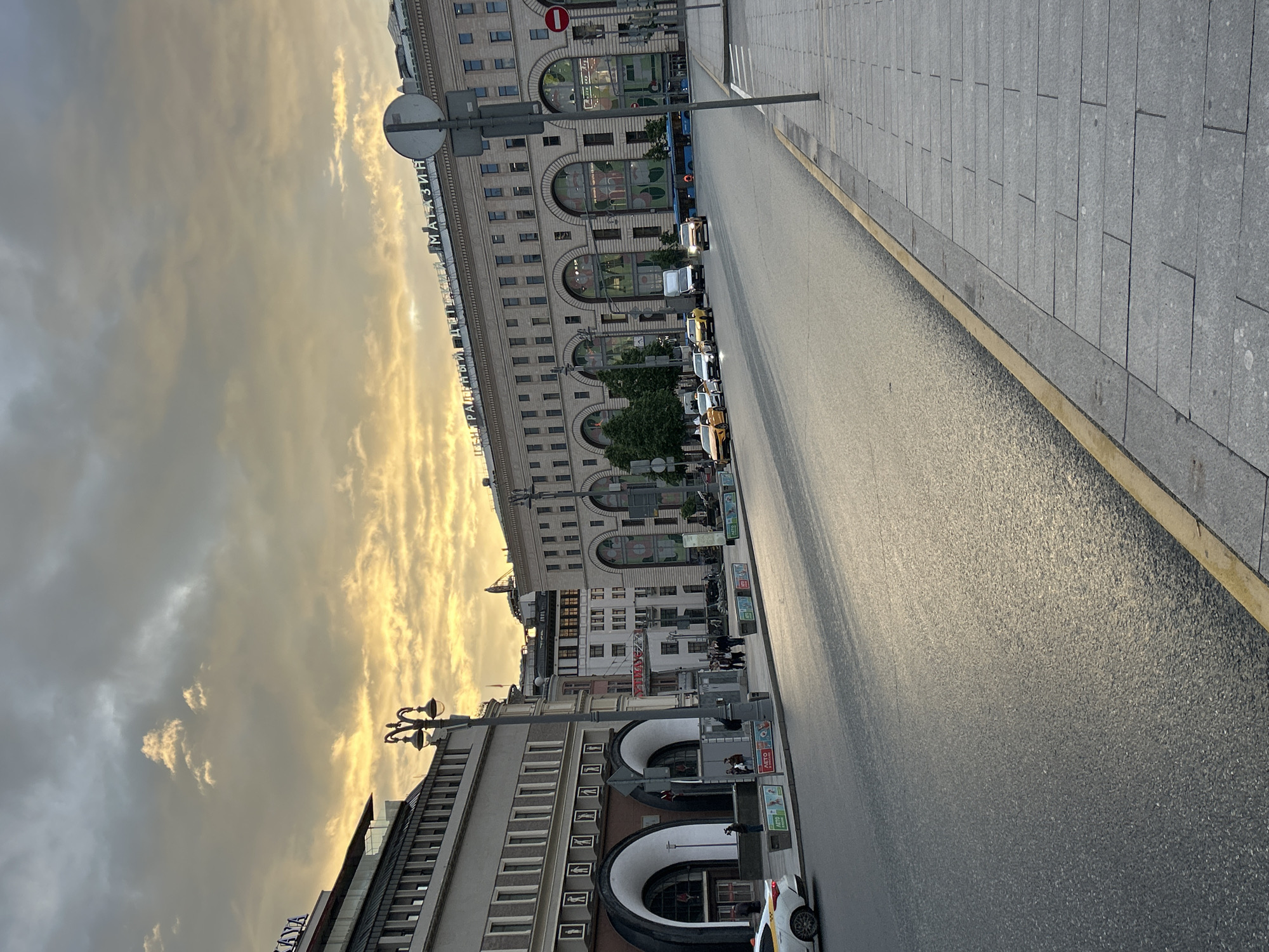 The image captures a street scene in an urban environment. The sky is filled with dramatic clouds, hinting at either sunrise or sunset, with a mix of grey and golden hues. The street appears relatively empty, with a smooth asphalt surface reflecting the sky's light.
On the right side of the street, there is a sidewalk paved with large rectangular stones, bordered by a yellow painted line. The buildings on the left are a mix of architectural styles, featuring arched windows and a combination of brick and stone facades. Some buildings are tall and imposing, while others are shorter and more modern.
Several vehicles, including cars and what appear to be buses, are parked along the side of the road in front of the buildings. There are pedestrians walking on the side walk in the distance. A street light is visible, and there is signage in Russian on one of the buildings.
