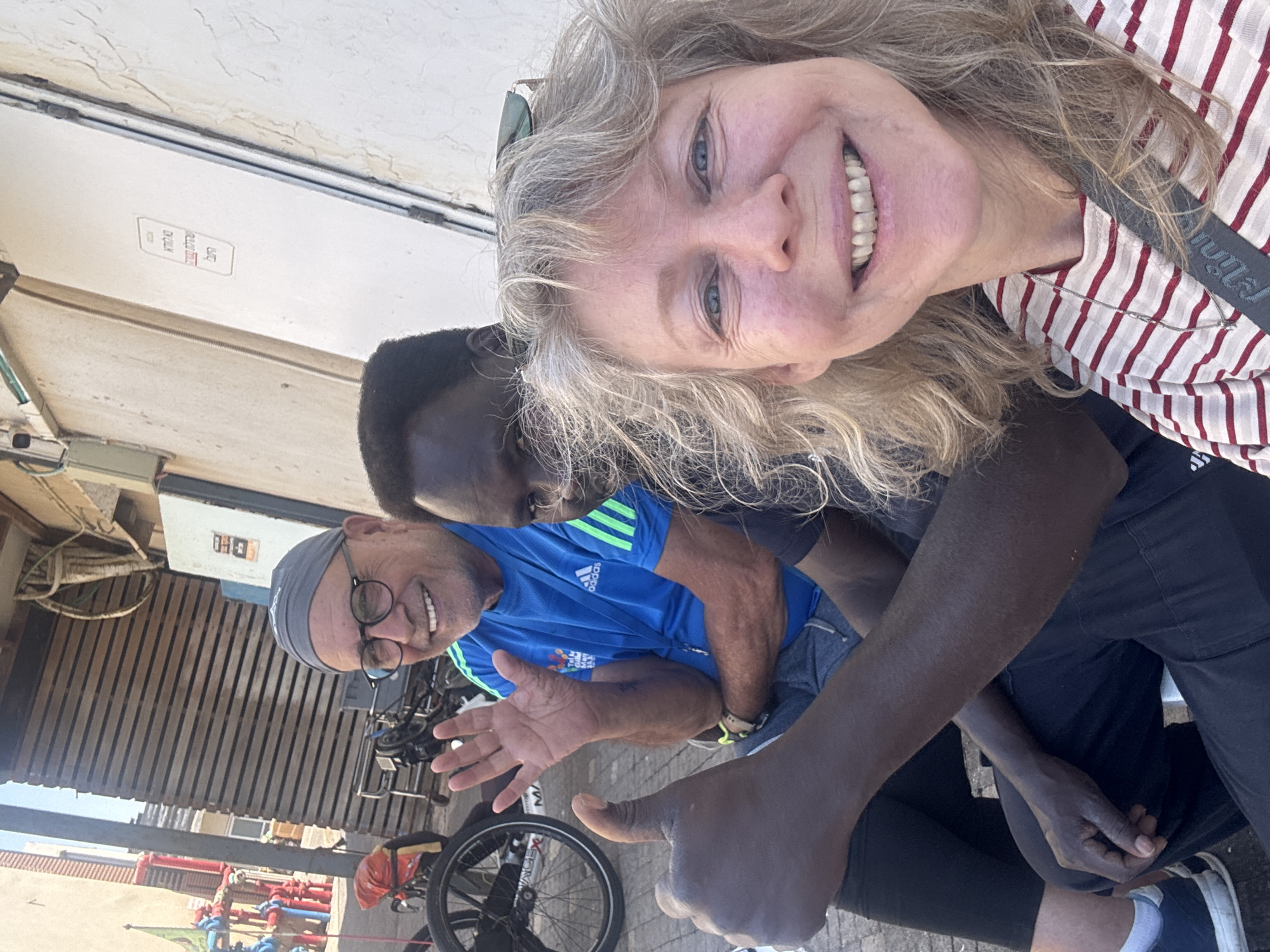 A selfie-style, medium-shot photograph shows four individuals outdoors. A woman with blond hair and a red and white striped shirt is positioned at the top, smiling broadly at the camera. Below her are three more figures. A man wearing a blue shirt with green stripes and a gray head covering is also smiling, his hand raised as if waving. Beside him is a person with dark skin who is partially obscured but looking towards the camera. Lastly, an arm extends into the frame, giving a "thumbs up" gesture.
In the background, there is a bicycle, various pipes painted red and green, and a slatted wooden structure. Above this structure is a white wall. The picture is taken outside in Tel Aviv, Israel.