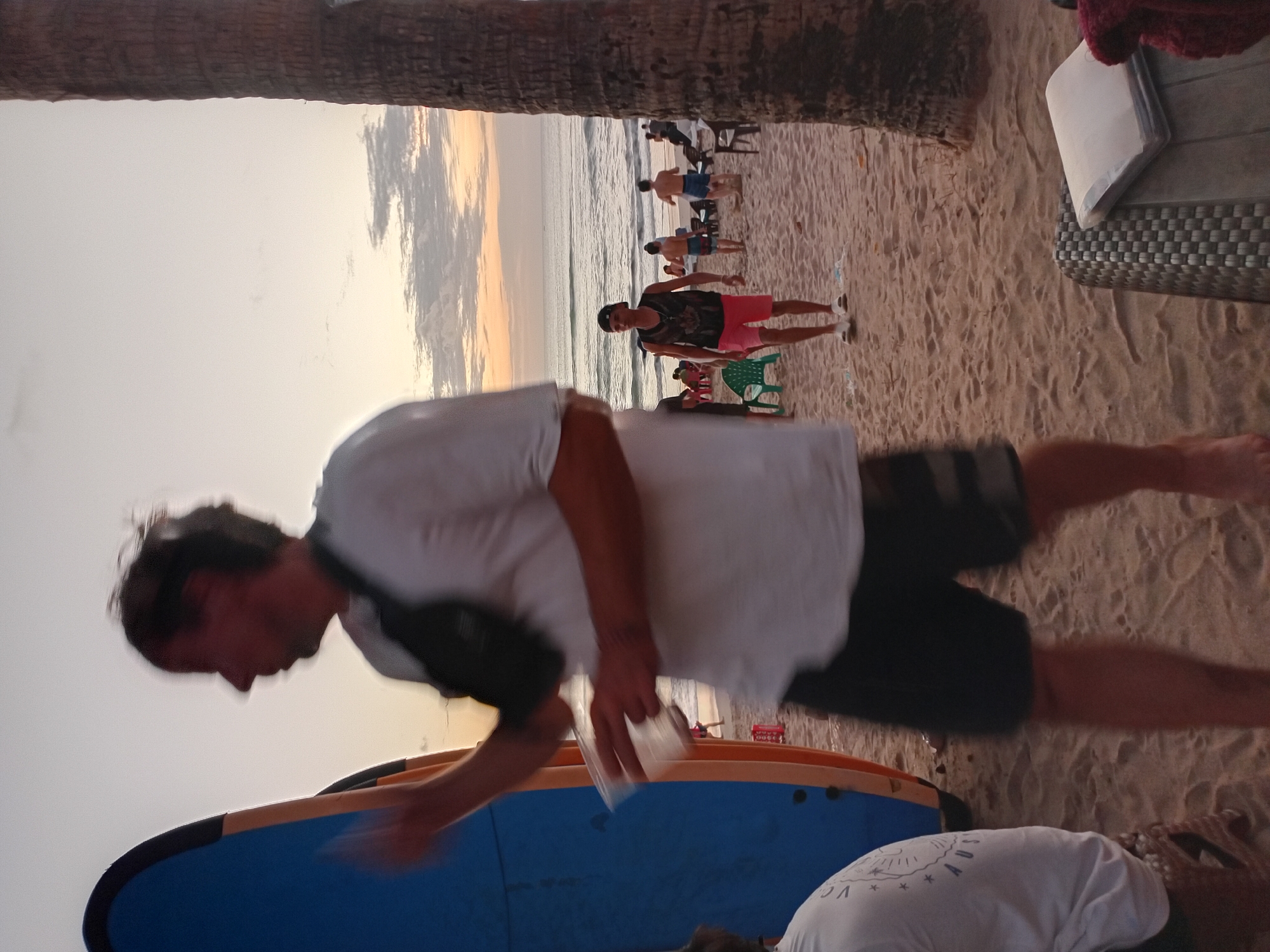A group of people are on a sandy beach in Kuta, Indonesia, near the ocean during sunset. In the foreground, a man in a white t-shirt and black shorts is walking away from the camera, with a blue surfboard to his left. He is holding a clear plastic cup in his right hand. Further back on the beach, a young man wearing a black sleeveless shirt with a design and pink shorts is walking towards the camera. Other individuals are scattered on the beach, some sitting in chairs and others walking near the water. Palm trees and the ocean are visible in the background. The sky displays a gradient of colors from the sunset.