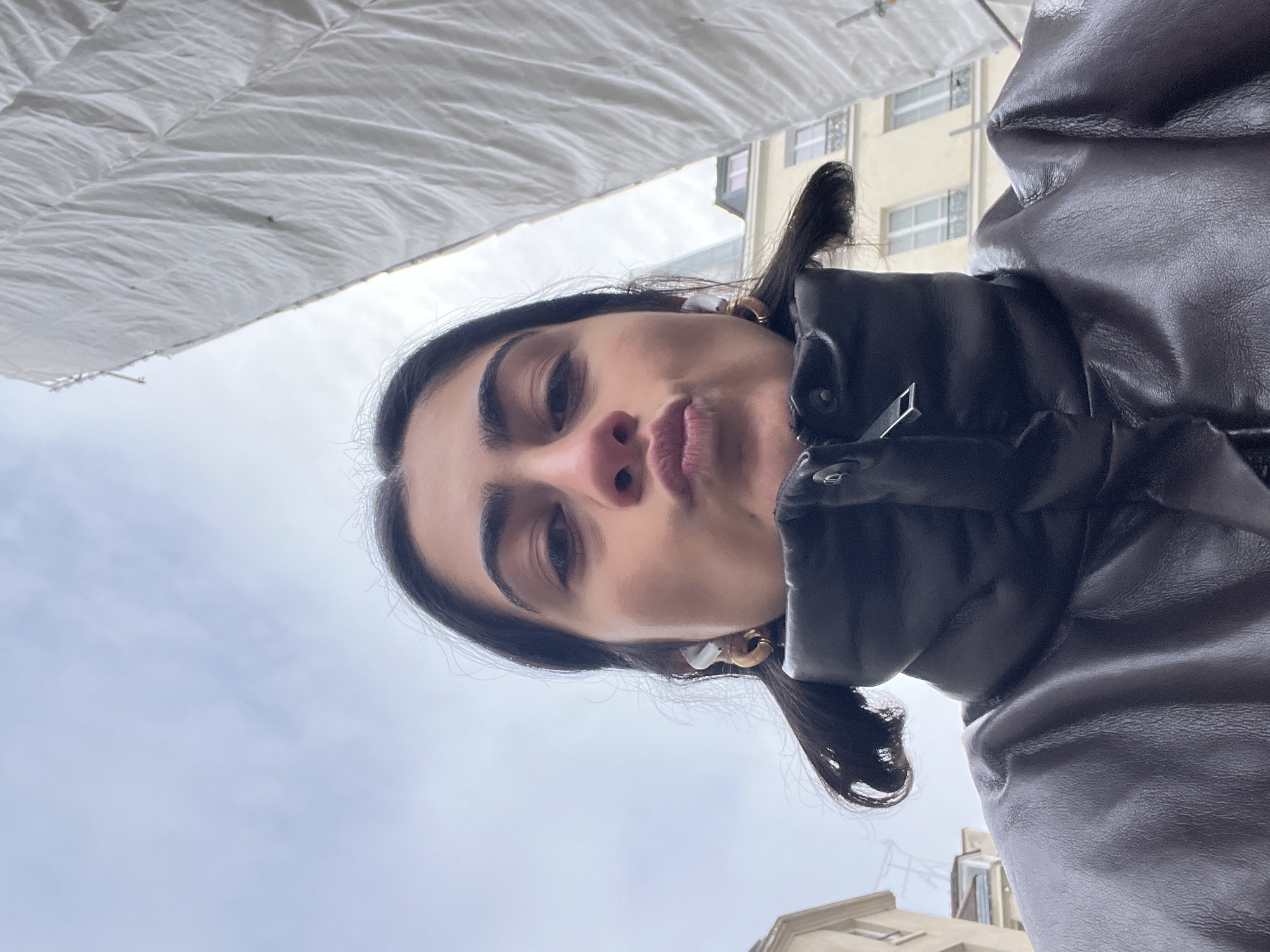 The image is a low-angle selfie of a young woman with dark hair, pulled into two pigtails, wearing a dark, possibly leather, puffer jacket. She has dark eyebrows and appears to be pursing her lips slightly. She is wearing gold hoop earrings and white earbuds.
The background includes a partly cloudy sky, rooftops of buildings, and a large white fabric canopy or awning above. The buildings visible have light-colored facades and windows. Given the architectural style and ambiance, the location appears to be in Paris, France.