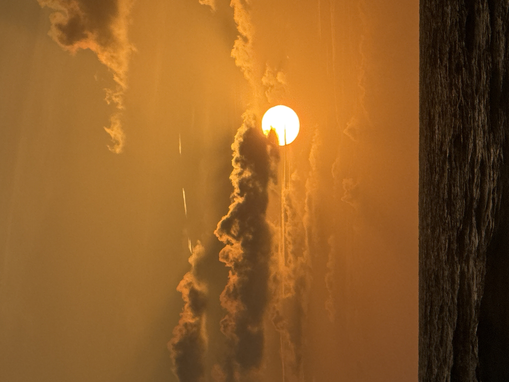 The image depicts a sunset over the ocean. The sun, a bright white orb partially obscured by clouds, is visible in the right-center of the frame. The sky is a vibrant orange, with wispy clouds scattered across it. The ocean in the foreground is dark and appears to be choppy, with visible waves. The horizon line is clearly defined, separating the sky from the sea. There are no people or man-made structures visible in the image. The location is identified as Karon, Thailand.