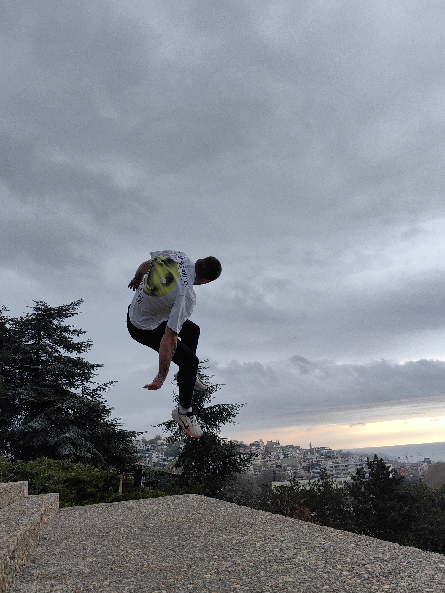 A man is captured mid-air, seemingly jumping from a concrete structure in Varna, Bulgaria. He is wearing a white t-shirt with a graphic on the back, black athletic tights, and athletic shoes. His body is angled downwards and his arms are extended. The background features a cloudy sky and a cityscape with buildings and trees. The scene appears to be taken from a high vantage point, overlooking the city.