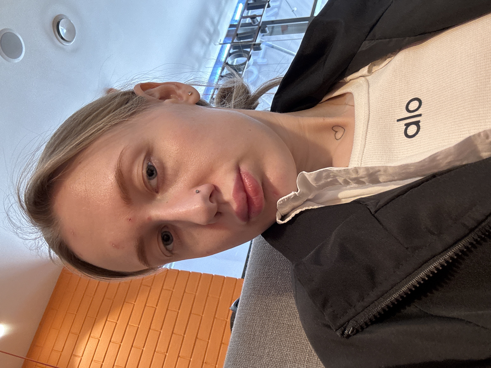 A selfie of a blonde woman wearing a white "alo" shirt with a black jacket. The woman has a nose piercing and a small heart tattoo on her neck. The background includes a blurred orange wall and a view through a window with what appears to be a metal structure outside. The image shows the woman from an unusual angle with her head tilted.
It is impossible to determine the location in the city of Rostov-on-Don, Russia from the image.