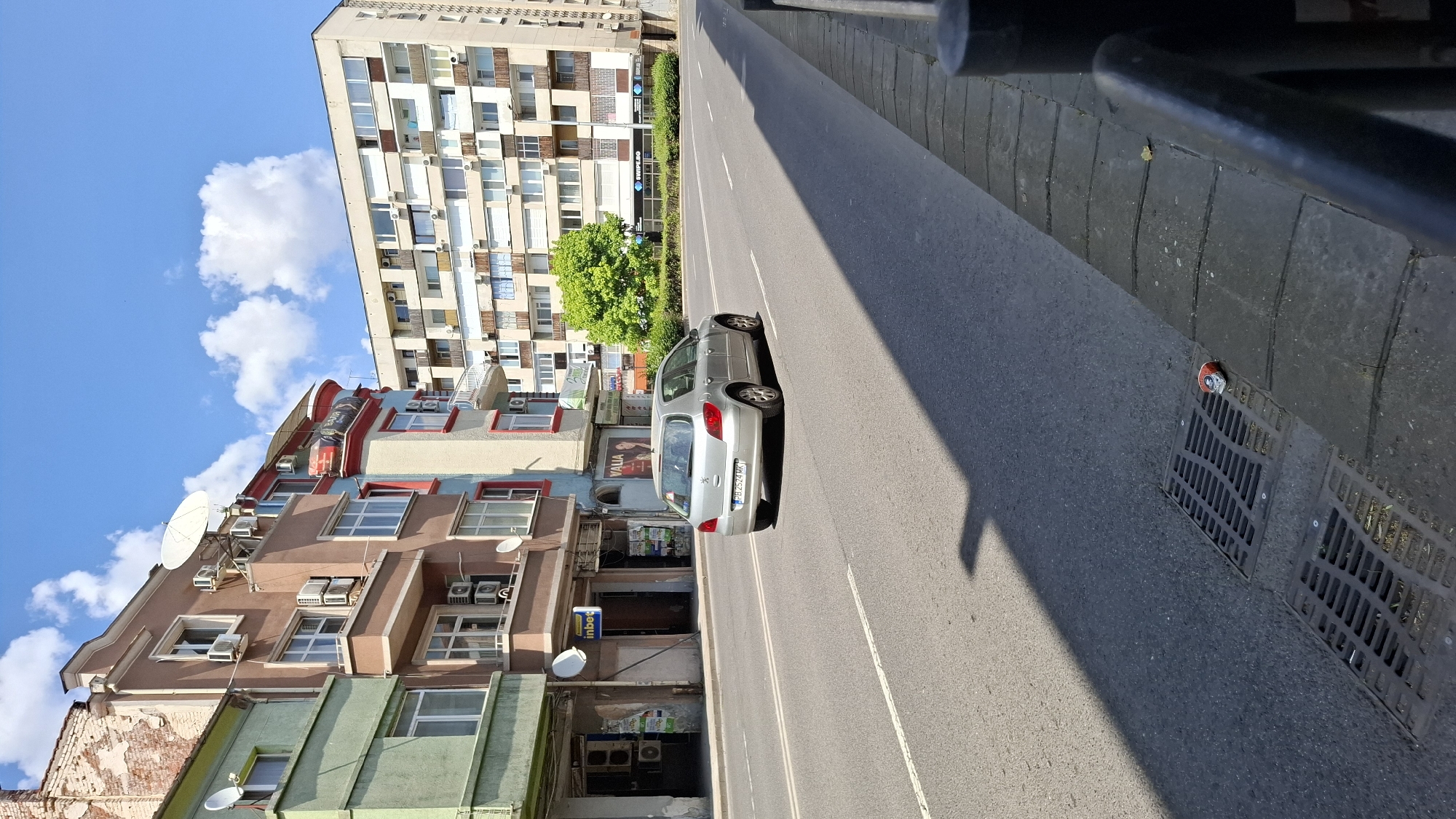 A silver Peugeot 307 hatchback, bearing Bulgarian license plate PB 2524 MK, drives on a multi-lane asphalt road in Plovdiv, Bulgaria. The vehicle is moving away from the camera, casting a shadow on the road. The road is bordered by a sidewalk with a metal grate and a black metal railing on the right. On the left side of the road are several multi-story buildings with a mix of architectural styles. The building closest to the camera on the left is painted green on the lower floors and appears older, with exposed brickwork on its upper section. Adjacent to it is a pinkish-brown building with balconies and numerous air conditioning units. Further down the road, a larger, beige apartment building with many windows is visible. A bright green tree stands in front of the beige building. The sky above is blue with scattered white clouds. The scene appears to be a street in an urban environment.