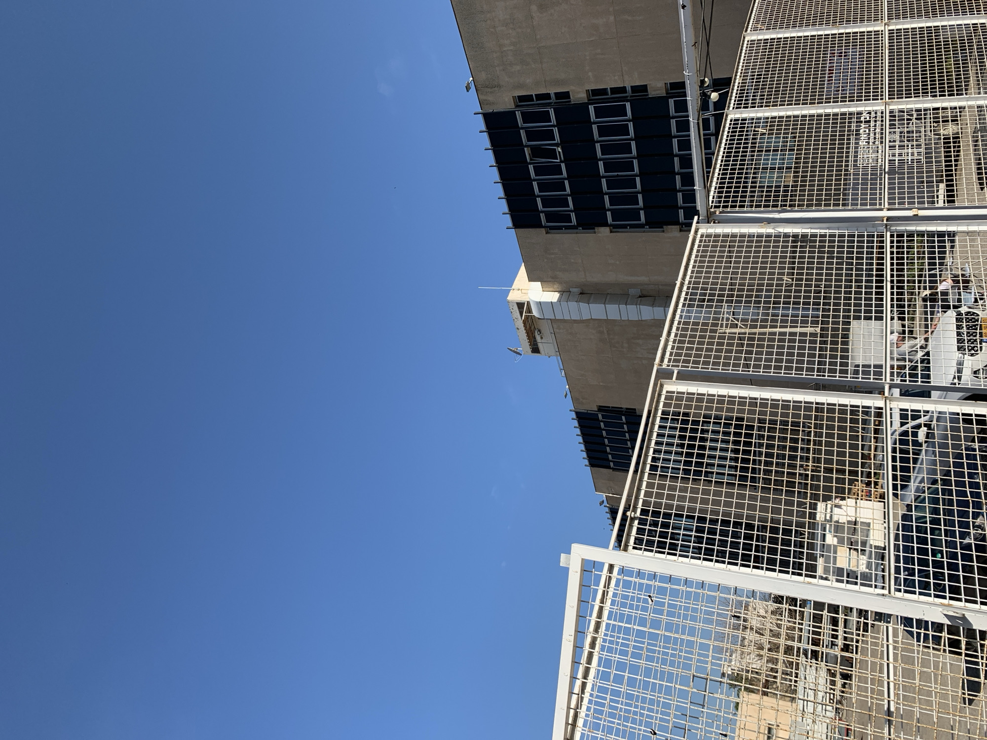 The image presents a daytime exterior scene in Tel-Aviv, Israel, featuring a multi-story building identified as part of the Ministry of Defense (Hakirya) complex. A white metal mesh security fence dominates the foreground, partially obscuring the view.
Behind the fence, multiple vehicles are visible, including a dark-colored car on the left and a white SUV or truck with a partial Israeli license plate reading "888803" in the center. Faint human figures are discernible near the white vehicle, suggesting activity in the enclosed area. A sign bearing the Hebrew text "פינוי בינוי" (Pinui Binui, meaning Urban Renewal) is visible behind the fence, indicating a construction or redevelopment zone.
The background features a large, utilitarian building with a light brown concrete facade. Its design incorporates prominent dark vertical panels containing multiple rows of white-framed rectangular windows, several of which are open. A large, light-colored external ventilation or duct system runs vertically up the left side of the building, connecting to a rooftop structure that includes a satellite dish and other communication equipment. The roofline is flat, featuring security lighting fixtures. The sky above is clear and uniformly blue.