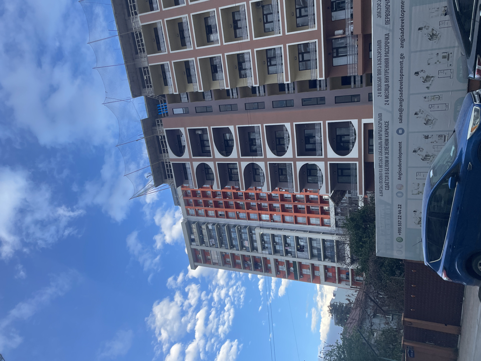 The image depicts a tall apartment building under construction in Batumi, Georgia, partially obscured by a promotional sign and a blue car. The building features multiple stories with balconies and a facade of brown, orange, and white sections. Construction netting is visible on the upper floors. The sky is blue with scattered white clouds. The promotional sign contains Georgian and Russian text, along with a phone number and email address associated with "Angisadevelopment."