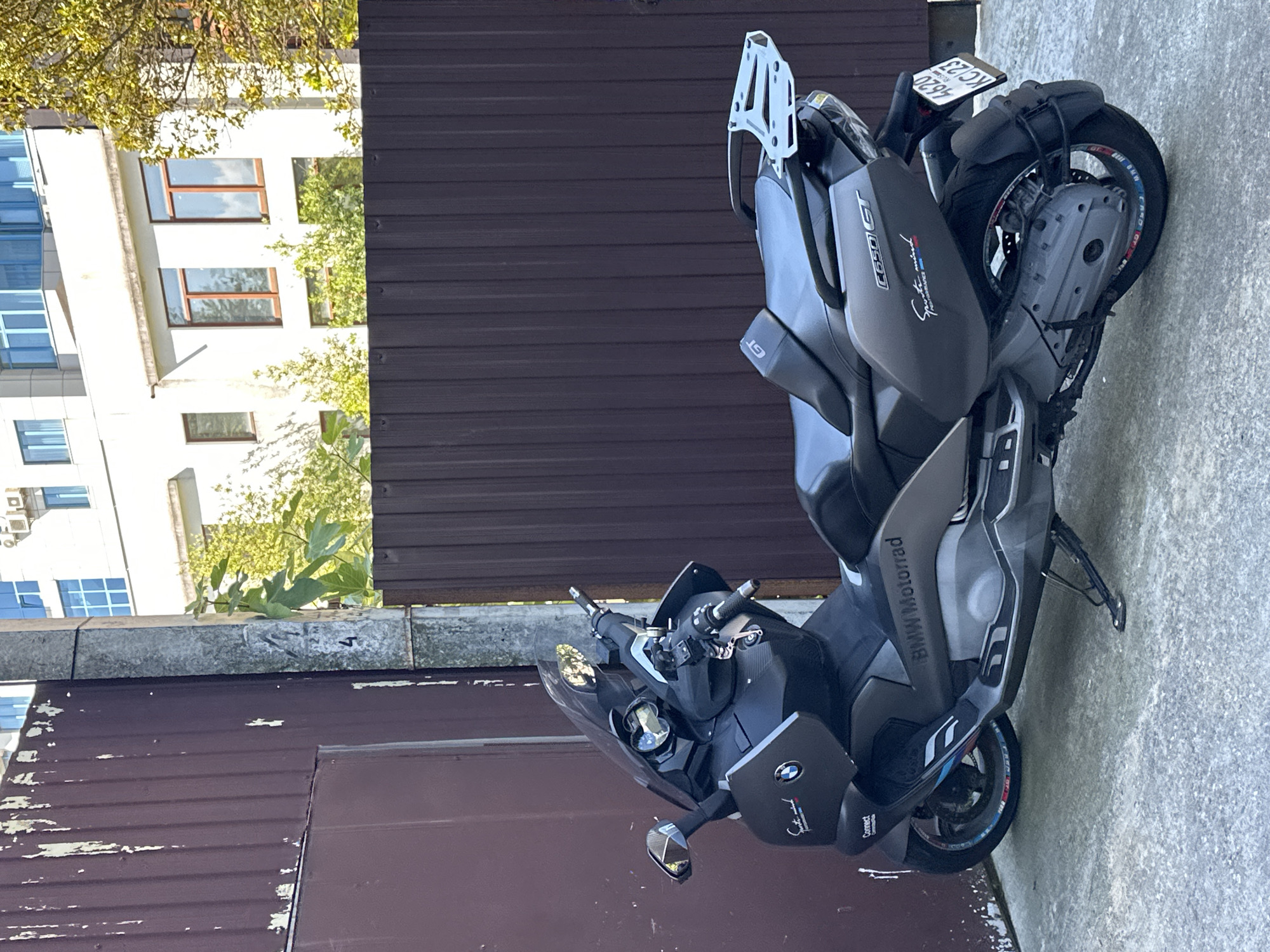 A dark gray BMW C650 GT scooter is parked on a concrete surface in front of a tall, corrugated brown metal fence. The fence is weathered and has peeling paint. Behind the fence, a modern white building with blue-trimmed windows is visible. Some trees with green foliage are also present. The scooter has a black seat, a clear windshield, and a silver rear rack. A license plate with the inscription "4620 KC 23" is attached to the rear of the scooter. The environment appears to be an urban setting, possibly a courtyard or alleyway. There are no people visible in the image. The lighting suggests it is daytime, likely outdoors. The image was taken in Sochi, Russia, as indicated by the license plate format.