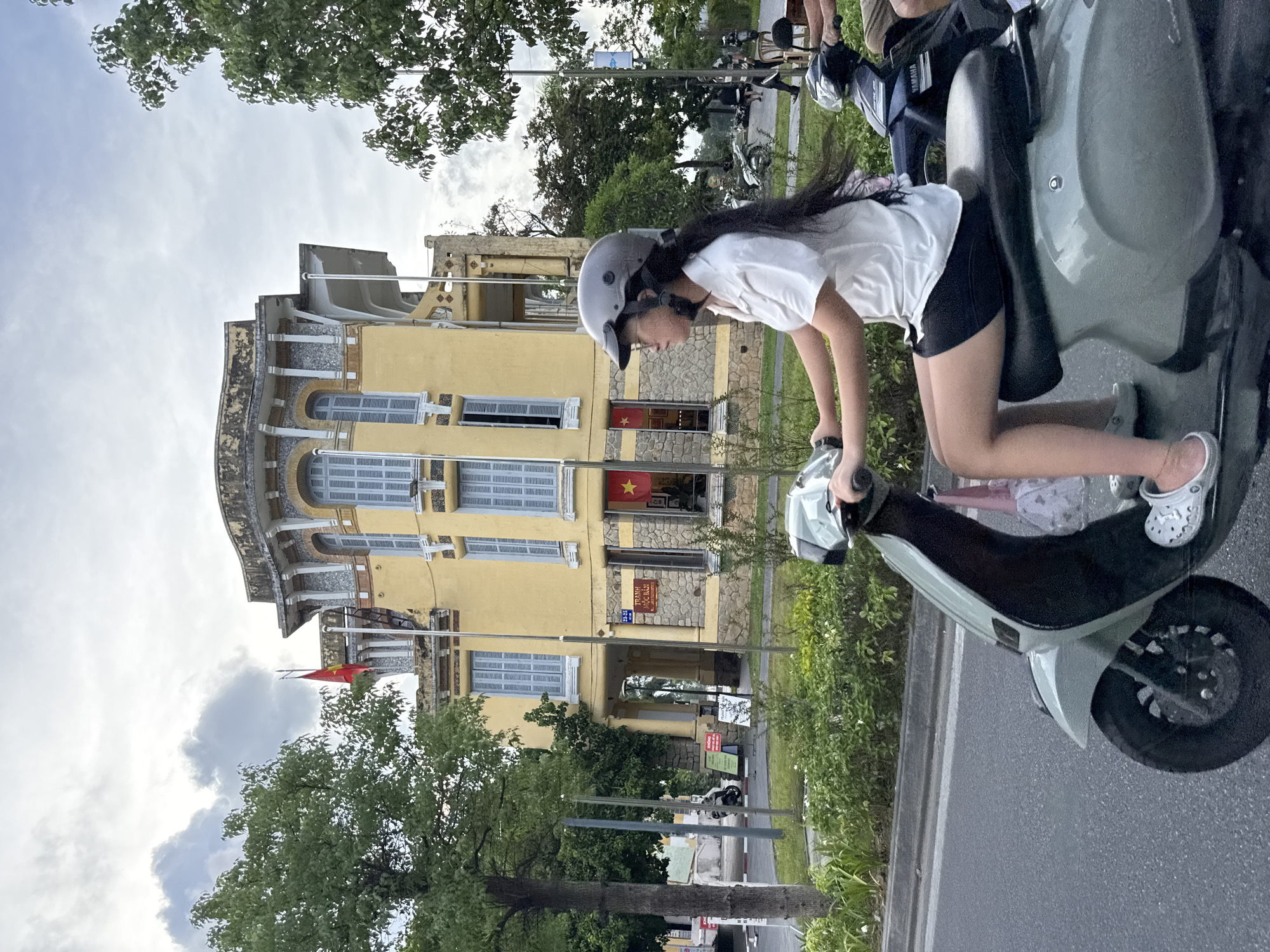 In Thuận Hóa Ward, Vietnam, a young Vietnamese girl wearing a helmet and a white shirt is riding a light green scooter. She has long dark hair and is wearing black shorts and white clogs. She is looking to her left. Behind her, a dark blue Yamaha scooter is partially visible. In the background, a large, old yellow building with ornate architectural details and light blue shutters stands prominently. A Vietnamese flag is flying on a flagpole in front of the building. The area is lined with trees and green foliage, and there is a street with other scooters and pedestrians in the distance. The sky is overcast with clouds.
