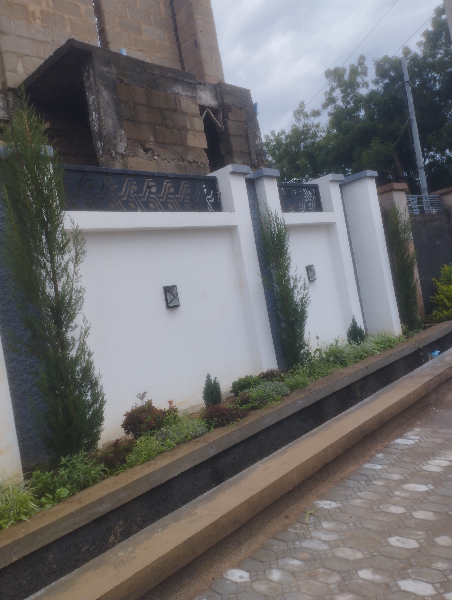 This image depicts a street scene in Jos, Nigeria, featuring a white plastered wall with a decorative metal railing. The wall is adorned with two square sconces, and slender evergreen trees are planted along its base. Beyond the wall, the top portion of an unfinished brick building is visible. In the foreground, a concrete structure, possibly a drainage channel, runs parallel to the street. The street itself is paved with interlocking hexagonal concrete blocks. The environment appears to be overcast, with trees visible in the background. No people are present in the image.