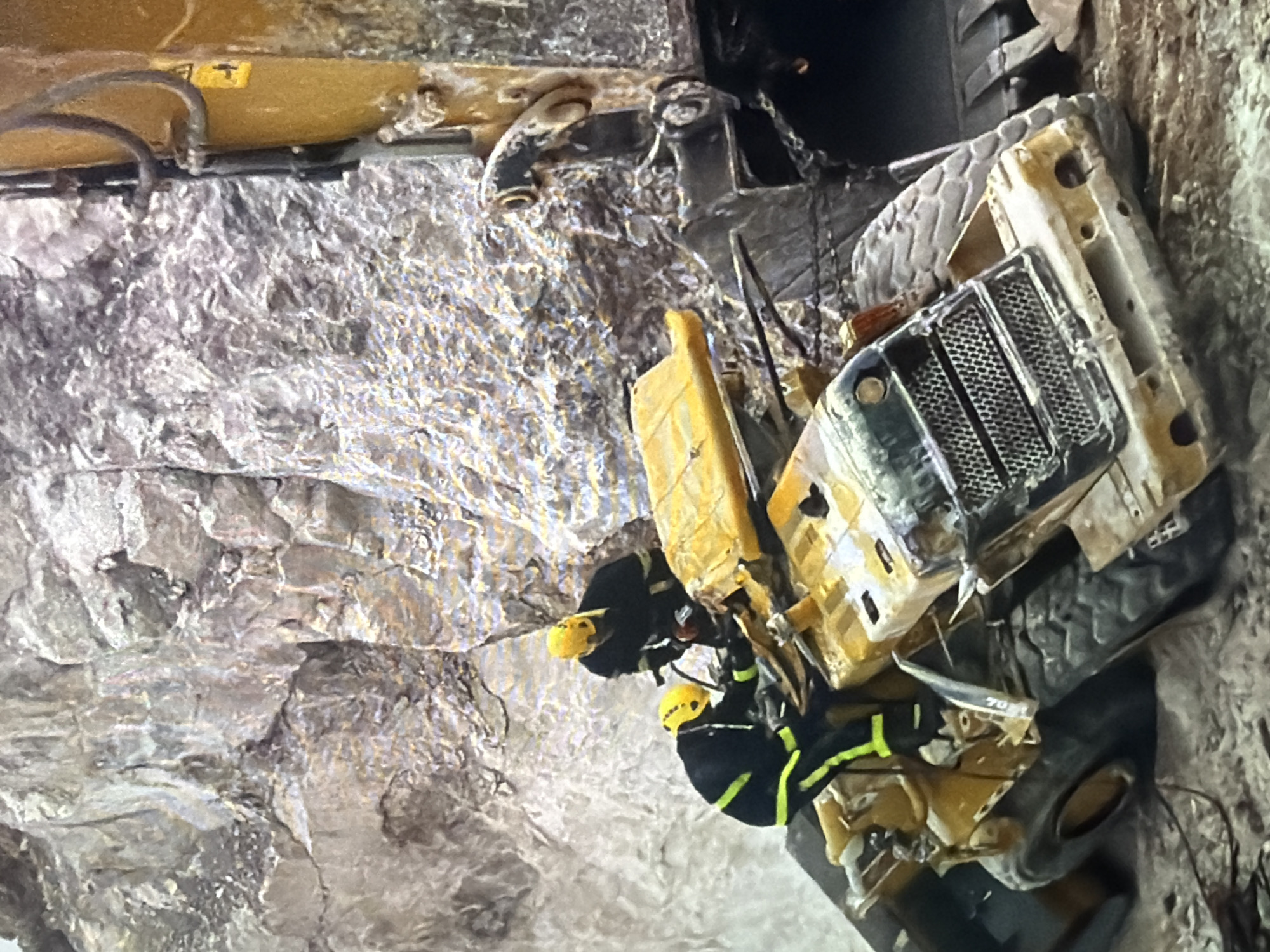 In an outdoor environment with a rocky, mountainous backdrop, two individuals in yellow helmets and black and neon-yellow safety suits are attending to a severely damaged yellow construction vehicle. The vehicle appears to be a front-end loader, overturned and partially buried in debris. The front grill is visible, as are large tires. One rescuer is positioned near the front of the vehicle, appearing to examine it, while the other is further back, leaning over the overturned machine. The overall scene suggests a rescue operation following a construction or mining accident. The location is not specified as being within a city.
