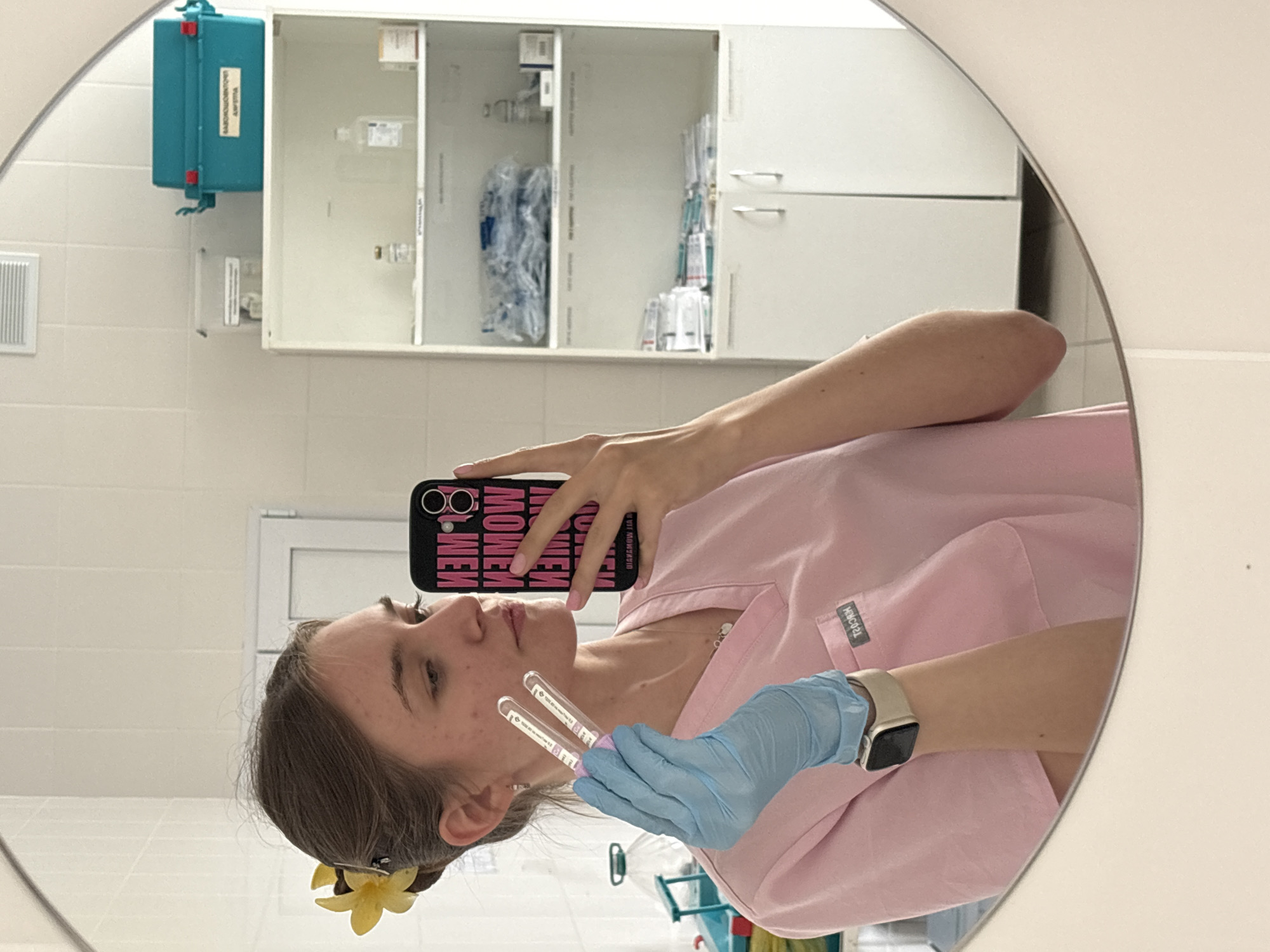 A woman in a light pink medical scrub suit and blue medical gloves is taking a selfie in a round mirror. She is holding two clear, sealed test tubes in her gloved hand. Her phone has a black case with pink lettering. She has a yellow flower in her hair. The background shows a medical setting with white tiled walls and white cabinets containing medical supplies. A blue box is mounted on the wall. The location is likely in the city of Perm, Russia, given the reflected writing on the phone case.