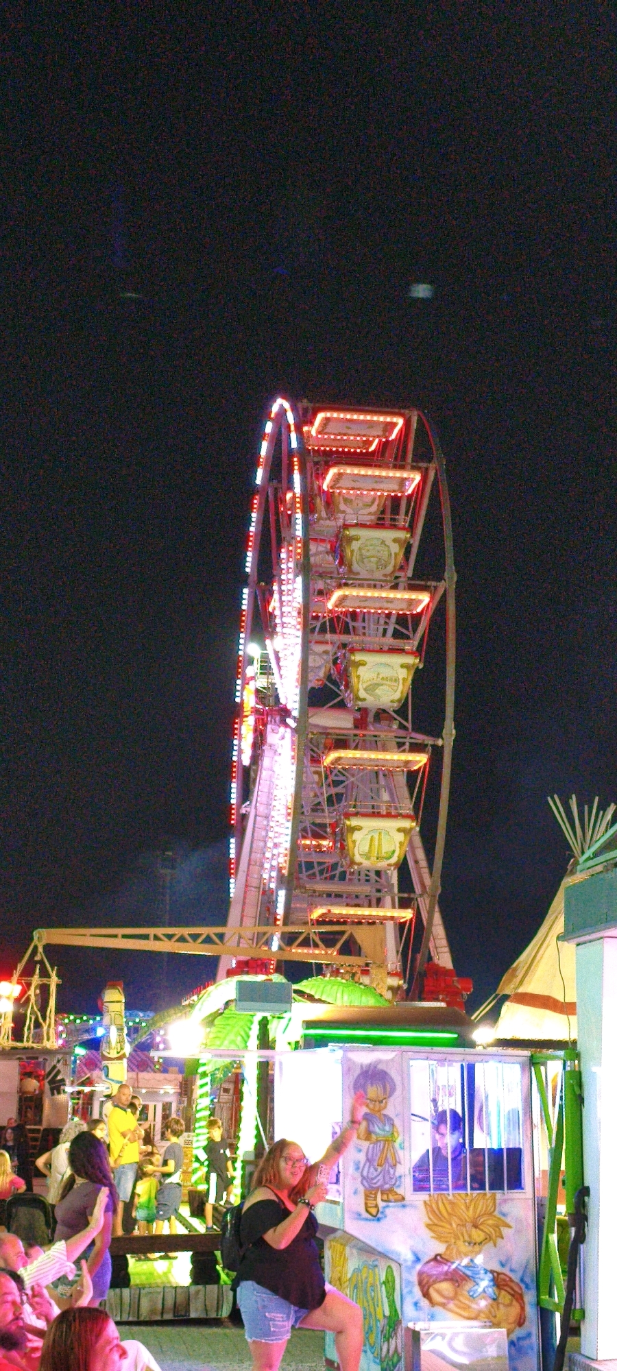This is a nighttime photograph of a fairground in Campiglia Marittima, Italy. In the foreground, a woman with glasses and a black off-the-shoulder top is holding up a phone, possibly taking a picture or recording. She is wearing denim shorts and a backpack. She is standing in front of a booth decorated with anime characters. To her left and behind her, other fairgoers are visible, some walking and some standing.
The middle ground is dominated by a brightly lit Ferris wheel, adorned with red and white lights. Several gondolas are visible on the wheel, some illuminated with yellow lights. Below the Ferris wheel, there are other fairground attractions with colorful lights. To the right of the Ferris wheel, a portion of a tent-like structure is visible. The background is dark, indicating it is nighttime.