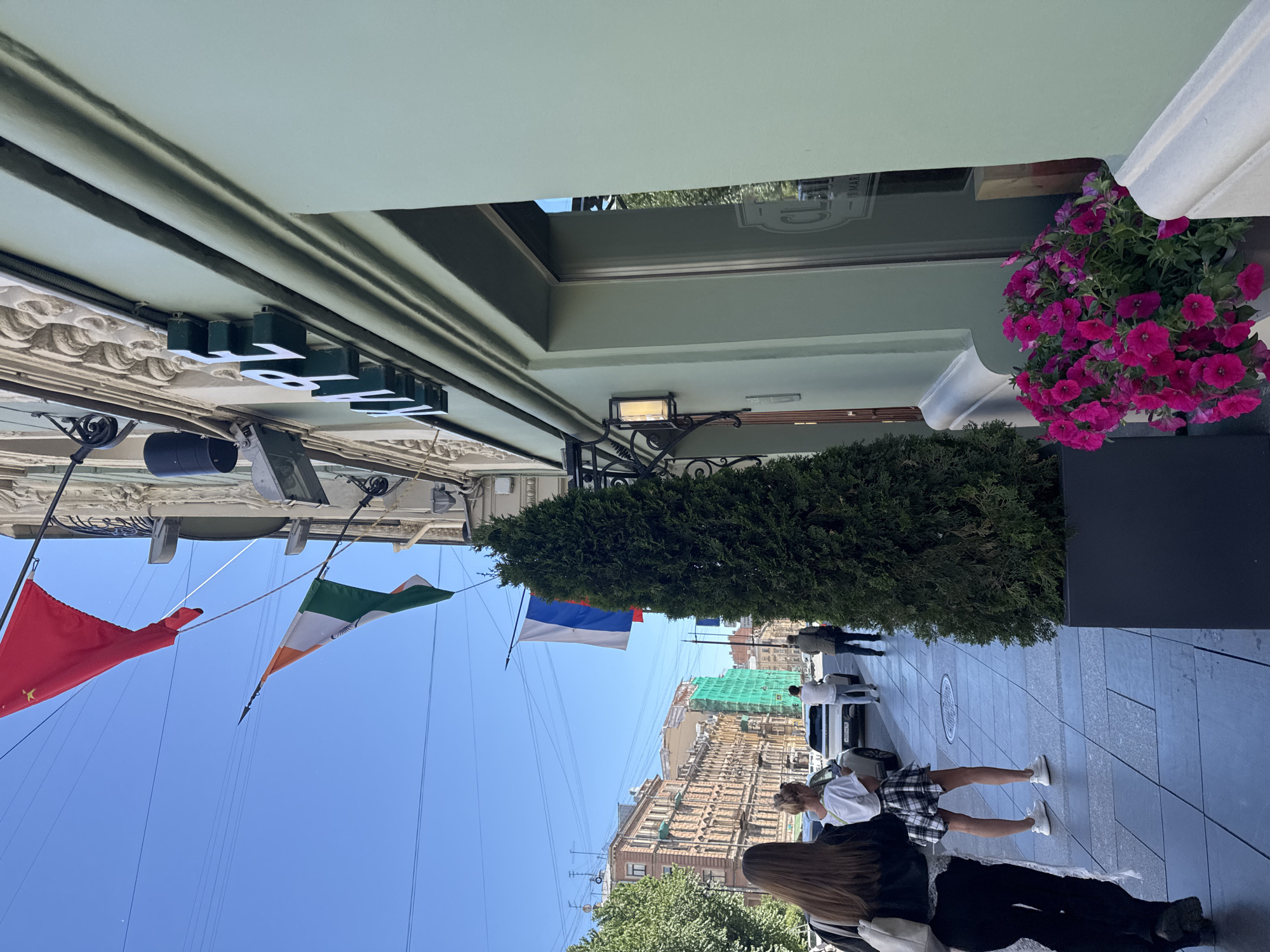 The image depicts a street scene in Saint Petersburg, Russia, on a clear, sunny day. The perspective is from a low angle, looking upwards at the building facades and the sky.
Prominently featured are several flags. A red flag, possibly the flag of China, is visible on the left. To its right, the flag of Ireland, with its green, white, and orange stripes, is displayed. Further along, the Russian flag, with its white, blue, and red stripes, is hung.
The architecture suggests an urban setting with ornate building details and decorative elements. A lush green shrub or small tree is planted in front of one of the buildings, providing a natural element to the urban landscape. In the background, several buildings are visible, some with scaffolding and construction nets, indicating ongoing development or renovation.
Several people are present on the sidewalk, walking in both directions. Their attire suggests a casual, possibly warm weather, day. Some individuals are walking alone, while others are in groups. One woman is walking with a plaid skirt and white top, and another person appears to be wearing a white shirt.
The image captures a slice of daily life in the city, with a mix of historical architecture, modern urban elements like construction, and the presence of flags suggesting a vibrant and international atmosphere. The lighting indicates it is daytime, likely late morning or early afternoon.