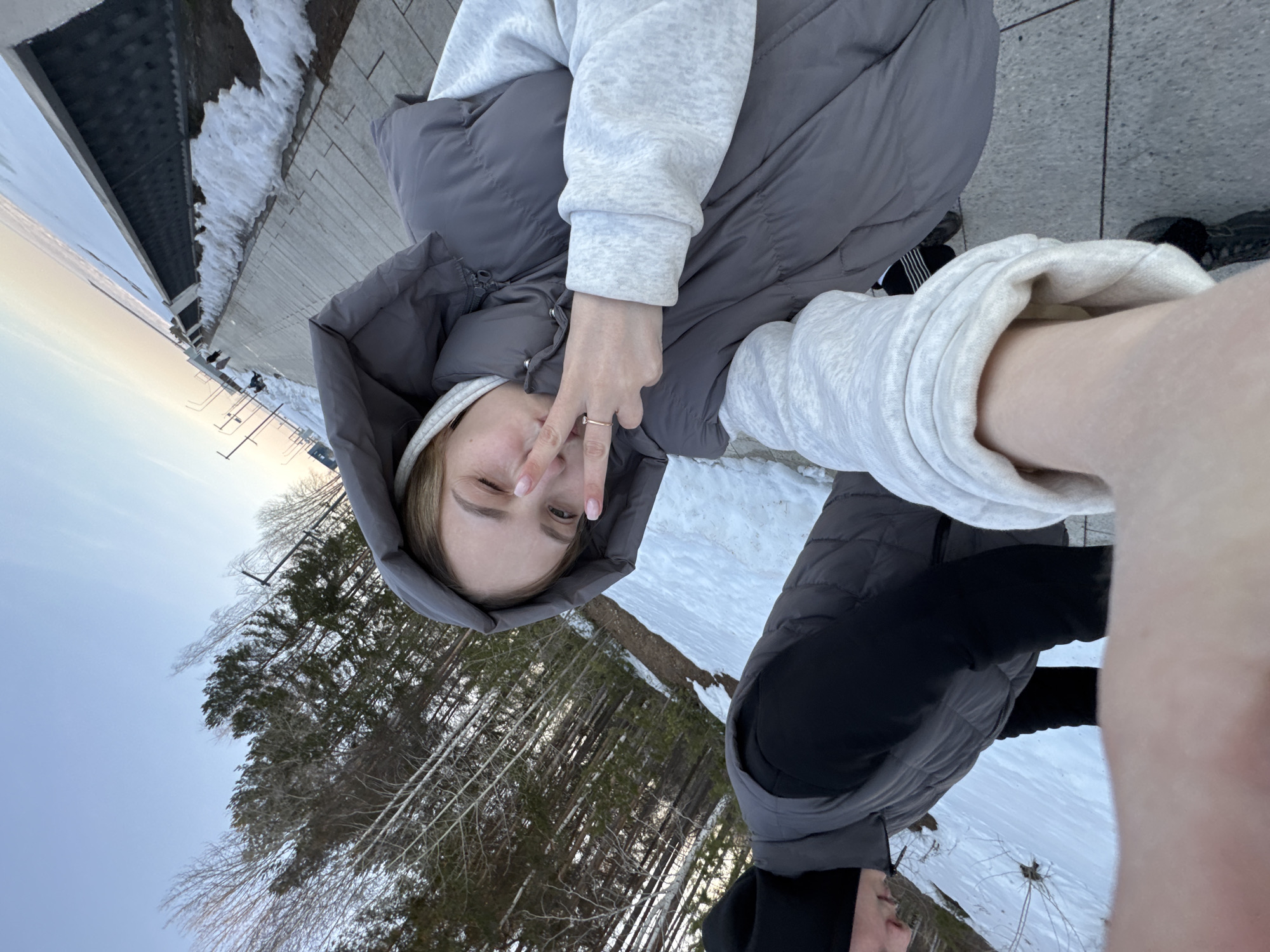 The image shows a selfie taken by a young woman outdoors in what appears to be a park or green space. The perspective is upside down. The woman is wearing a gray puffer vest with a hood, under which a light-colored beanie is visible. She is also wearing a light gray sweater. Her right hand is raised, with two fingers held up in a "peace" sign near her winking eye. She has light skin and light-colored hair.
Behind her, tall pine trees can be seen against a blue sky. The ground is partially covered in snow. To the left of the image, there is a view of a body of water in the distance and a walkway lined with snow and structures. The location is identified as the city of Chaikovsky, Russia, based on the trees and type of winter.
Another person partially appears in the lower right corner, also wearing a gray puffer vest and black clothing. This suggests that the young woman is not alone.