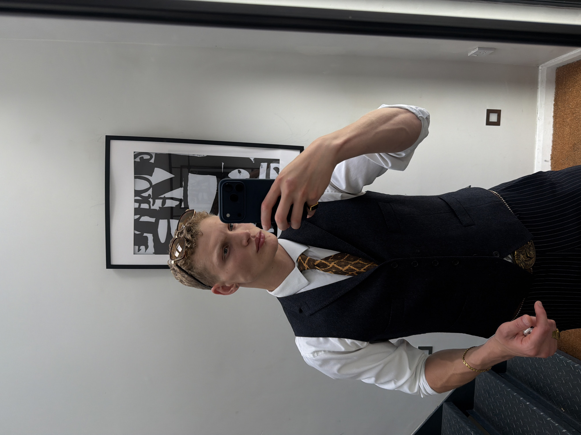 A young man with blond curly hair is taking a selfie in a mirror. He is wearing a white dress shirt, a dark vest, and striped trousers. He has sunglasses perched on his head and is wearing a gold bracelet and ring. Behind him, a framed piece of abstract art hangs on a white wall. The scene appears to be indoors, possibly in a residential setting, with a staircase visible to the left and a brown rug on the floor. There is no indication of the location being in London, United Kingdom.