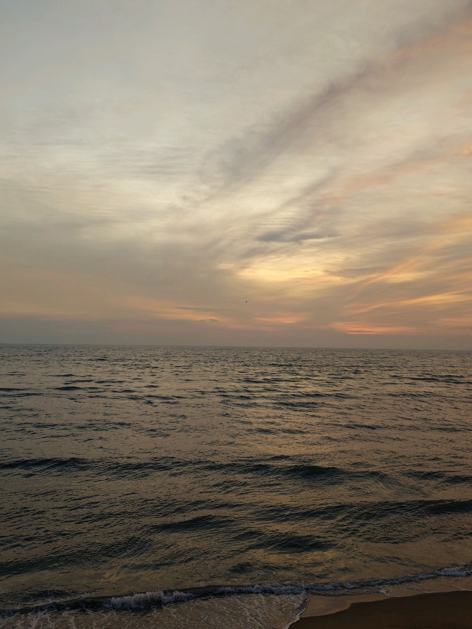 The image depicts a serene seascape at Anangadi, India, during what appears to be dusk or dawn. The horizon is marked by a calm expanse of water, with gentle ripples reflecting the soft light of the sky. The sky itself is a canvas of muted colors, featuring a gradient of pale yellow, orange, and hints of grey, indicative of the transition between day and night. Wispy clouds stretch across the sky, adding texture and depth to the scene. The lower portion of the image shows a narrow strip of sandy beach, with a delicate wave lapping at the shore, creating a foamy edge. There are no people or man-made structures visible in the frame, emphasizing the natural beauty and tranquility of the location. The overall atmosphere is one of peace and quietude.