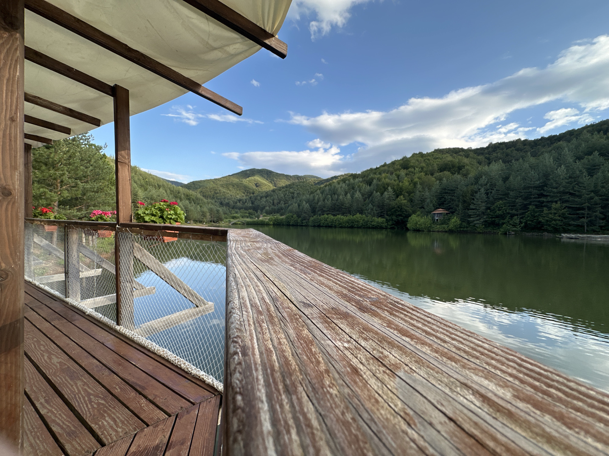The image shows a waterfront view from a wooden deck. The deck has a wooden railing with mesh fencing and a white rope trim. There are flower pots with red and pink flowers on the deck. In the background, a calm lake is surrounded by green, forested hills under a blue sky with white clouds. A small, rustic building with a red roof is visible nestled amongst the trees on the hillside. The scene suggests a natural, tranquil environment, possibly a resort or private property located near Kremen, Bulgaria. The lighting indicates it is daytime, likely late afternoon given the soft light on the landscape.