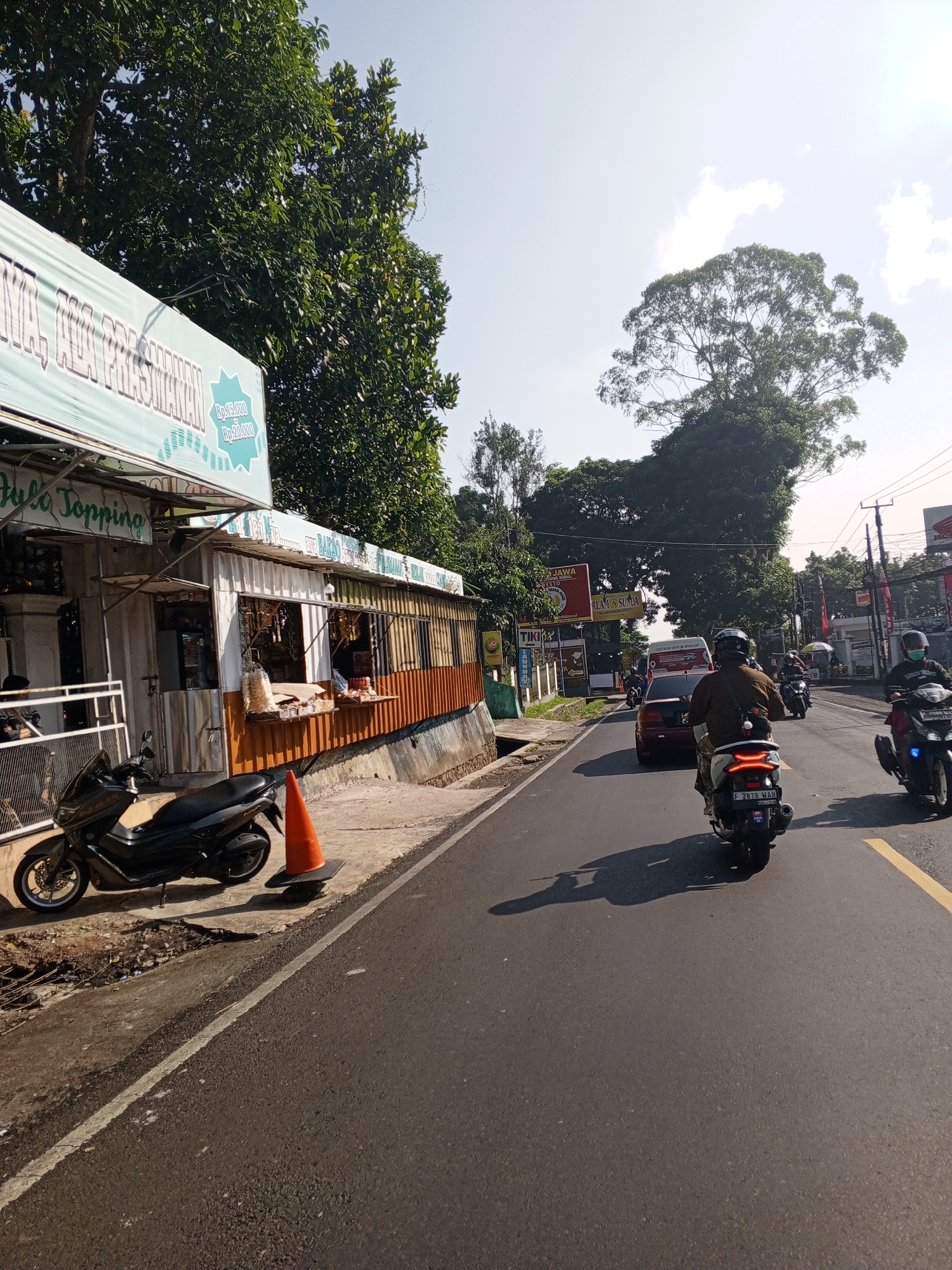 This image depicts a sunlit street scene in Weddagala, Sri Lanka, characterized by a mix of commercial establishments, dense vegetation, and ongoing road traffic. The time of day appears to be late morning or early afternoon, indicated by the clear blue sky with sparse white clouds and the distinct, elongated shadows cast by vehicles on the asphalt. On the left side of the frame, a single-story commercial building, possibly constructed from corrugated metal or similar materials, features an open storefront. An awning above the shop entrance displays a light blue banner with white text, partially legible as "Hulo Topping" and "LA PRAWANAN". Another section of the banner shows pricing in Indonesian Rupiah: "Rp.15.000" and "Rp.20.000". Goods, likely packaged snacks, are displayed on shelves within the open shop window. A person is visible inside the shop, possibly attending to customers. Parked on the concrete sidewalk in front of this shop is a black Yamaha NMAX scooter, facing left. An orange traffic cone is positioned on the sidewalk near the scooter. Further down the street on the left, additional signs are visible, including one with the text "ALAM SUNDA", and dense green foliage lines the roadside. The asphalt road, which appears dry, occupies the mid-ground and extends into the distance. It features a solid white line marking the edge and a broken yellow line delineating lanes. Traffic flows on the right side of the road. In the immediate foreground on the road, a person riding a white scooter is seen from behind, wearing a brown jacket and a dark helmet. The scooter's license plate, "F 2878 WAH," is clearly visible. To the right of this scooter, another person on a black scooter is also seen from behind, wearing a dark jacket, a dark helmet, and a face mask. Its license plate is partially visible as "D 6165 MAJ." Further up the road, a dark red sedan is visible behind the white scooter, and a white van or bus is partially seen behind the car. Several other motorcycles and distant vehicles are also present. The right side of the image is dominated by lush, green trees and vegetation. A particularly large, mature tree with a broad canopy stands prominently against the bright sky in the upper right quadrant. Utility poles with overhead wires run along the right side of the road. In the distance, various commercial signs, including one for "Honda," are discernible