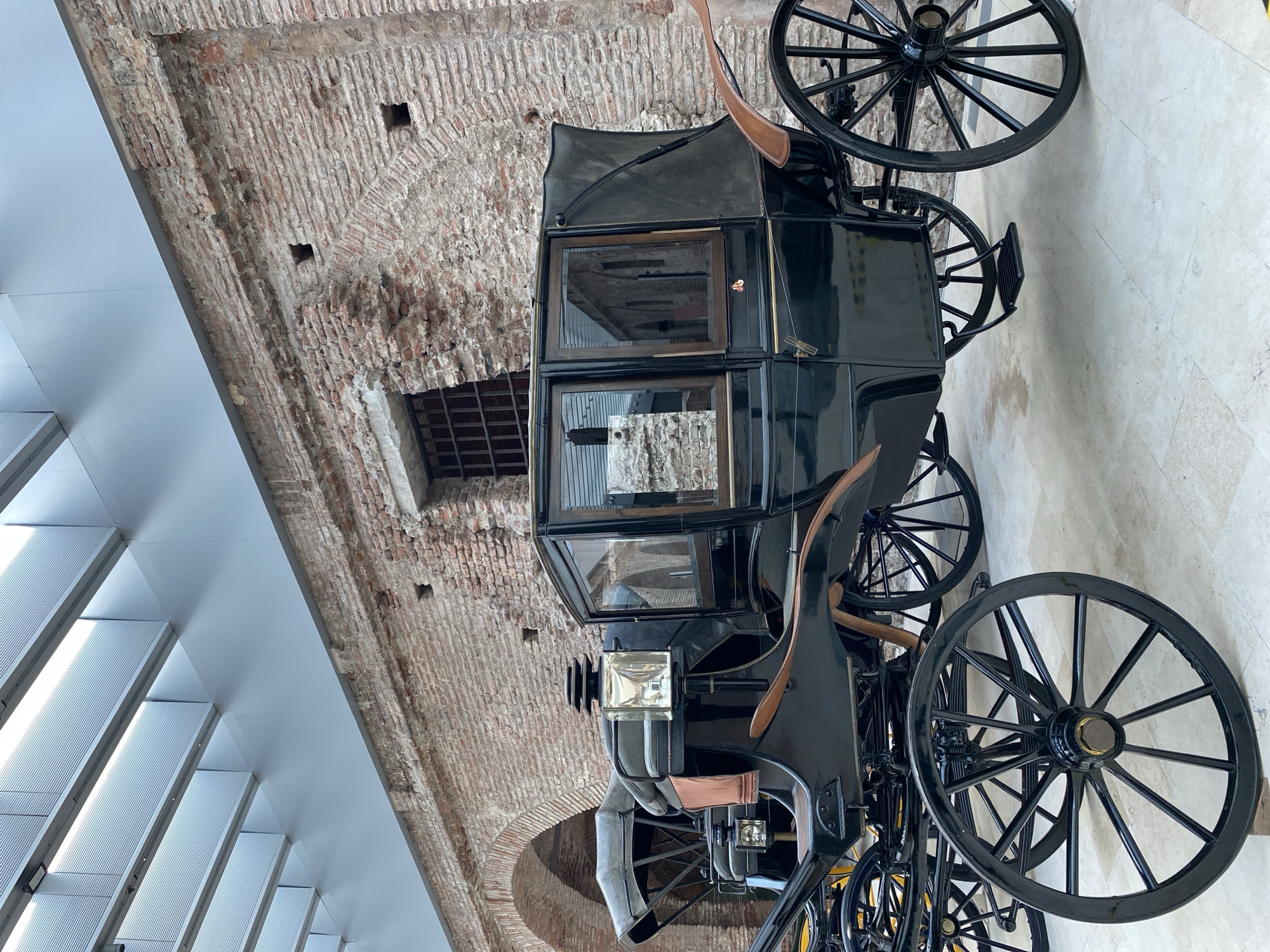 A large, antique, black horse-drawn coach or phaeton is the central subject of this image, displayed indoors, likely within a museum or exhibition space in Buenos Aires, Argentina. The carriage features a polished, dark chassis with prominent, spoked wheels painted black. The cabin is enclosed, with multiple clear glass windows framed in dark wood, and a black, possibly leather or canvas, roof. Two vintage-style lamps with reflective brass or metal casings are visible at the front.
The setting combines historical and modern architectural elements. Behind the carriage, a rustic, exposed brick wall with irregular patterns and mortar joints forms a significant part of the background. This wall shows signs of age, with some areas appearing worn or recessed, and features a small, barred opening or window. Above, a contemporary white ceiling structure with parallel, ridged panels, possibly skylights or integrated lighting, contrasts sharply with the ancient brickwork. The floor beneath the carriage is a light-colored, tiled surface.
The image captures the carriage in a static display, with no people present or activity taking place. The lighting suggests a well-illuminated indoor environment. There is no visible text on the carriage or in its immediate surroundings.