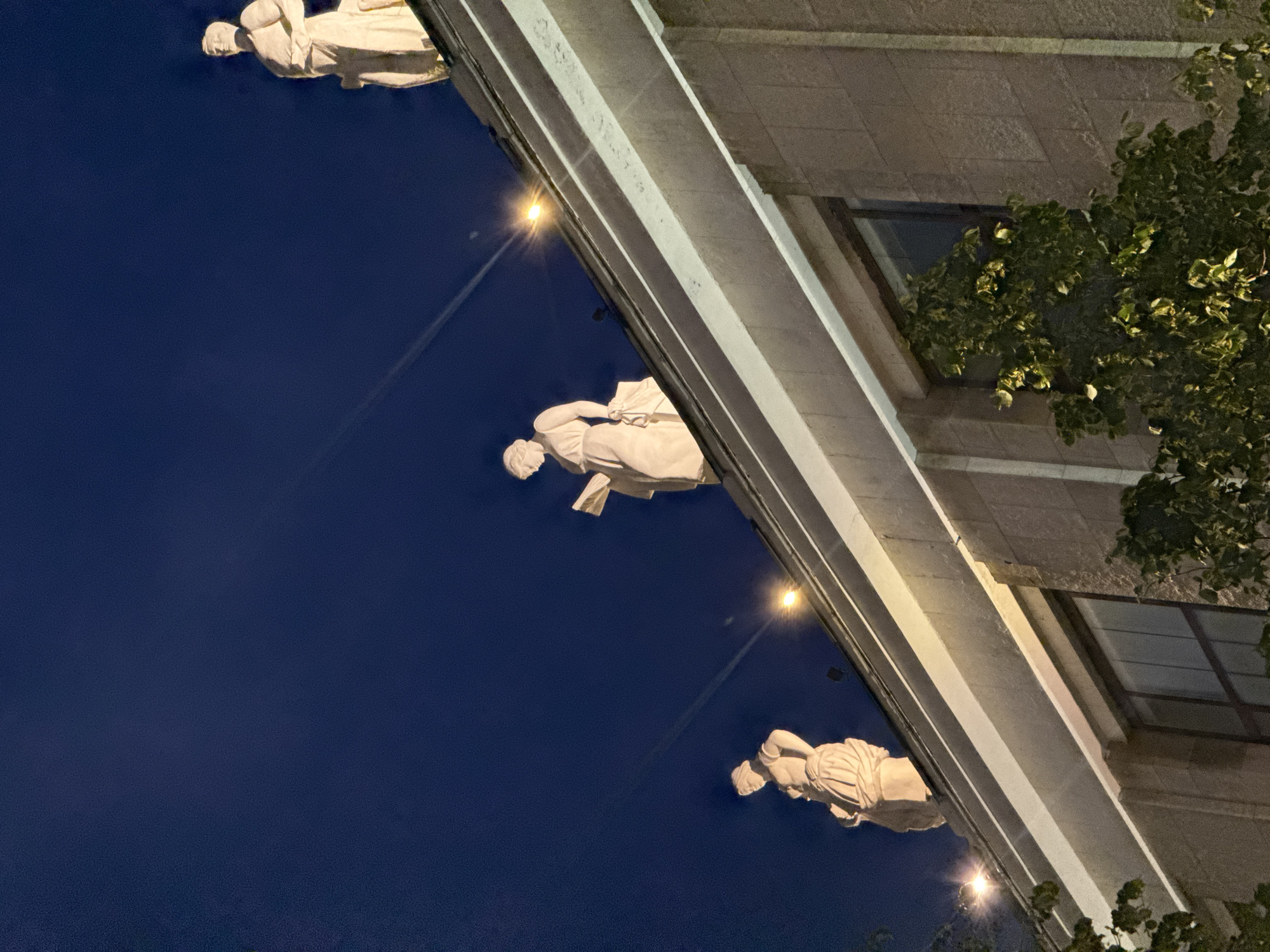 The image shows the facade of a building at night, with three illuminated classical statues perched on the rooftop. The statues, appearing to be made of light-colored stone, depict female figures in draped garments. The statue on the far left is slightly turned to the left. The middle statue is facing forward and appears to be reading a book. The statue on the far right is also facing forward. The building's facade is constructed of light-colored stone blocks, and a large window is visible on the lower right. Trees with green foliage are in the foreground, partially obscuring the lower portion of the building. The sky is a deep, dark blue, illuminated by bright lights from the statues and streetlights. This building is likely the Ministry of Foreign Affairs of the Russian Federation, located on Smolenskaya Square in Moscow, Russia.