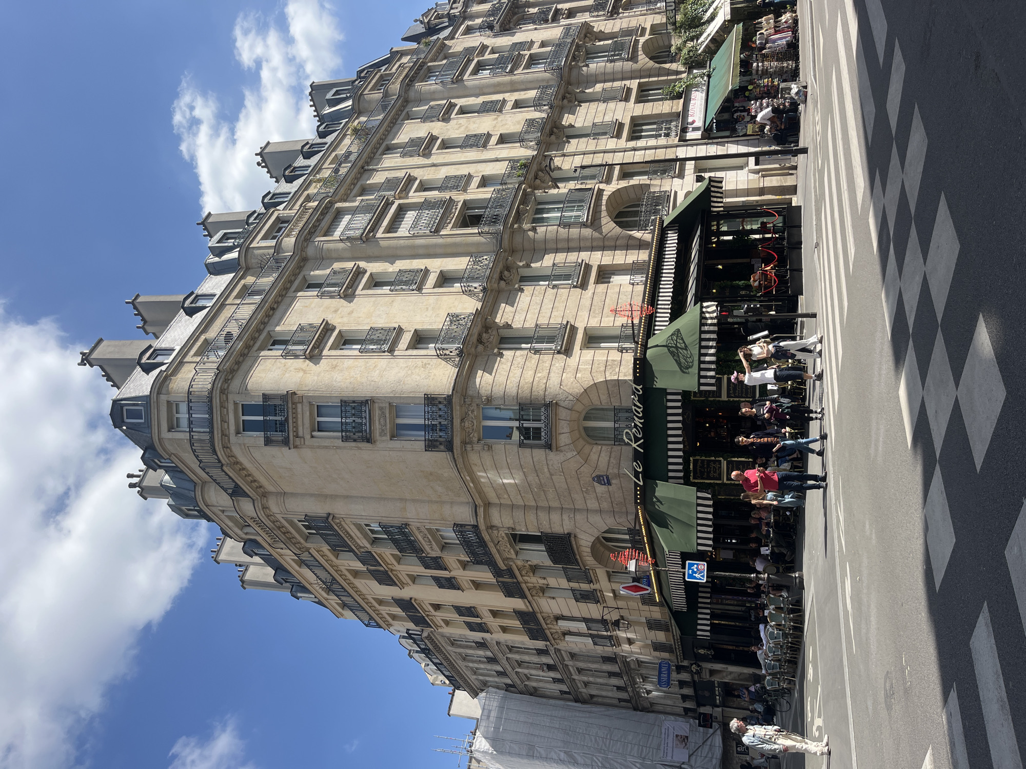 The image shows a street scene in Paris, France. The architecture consists of a tall, ornate building with multiple floors, a corner location, and balconies. The building is light-colored stone. A restaurant or cafe with a striped awning, named "Le Renard," is visible at street level. People are walking on the sidewalk and in a crosswalk. The crosswalk is marked with black and white stripes. The sky is blue with scattered clouds. The angle of the photo suggests it was taken from street level.