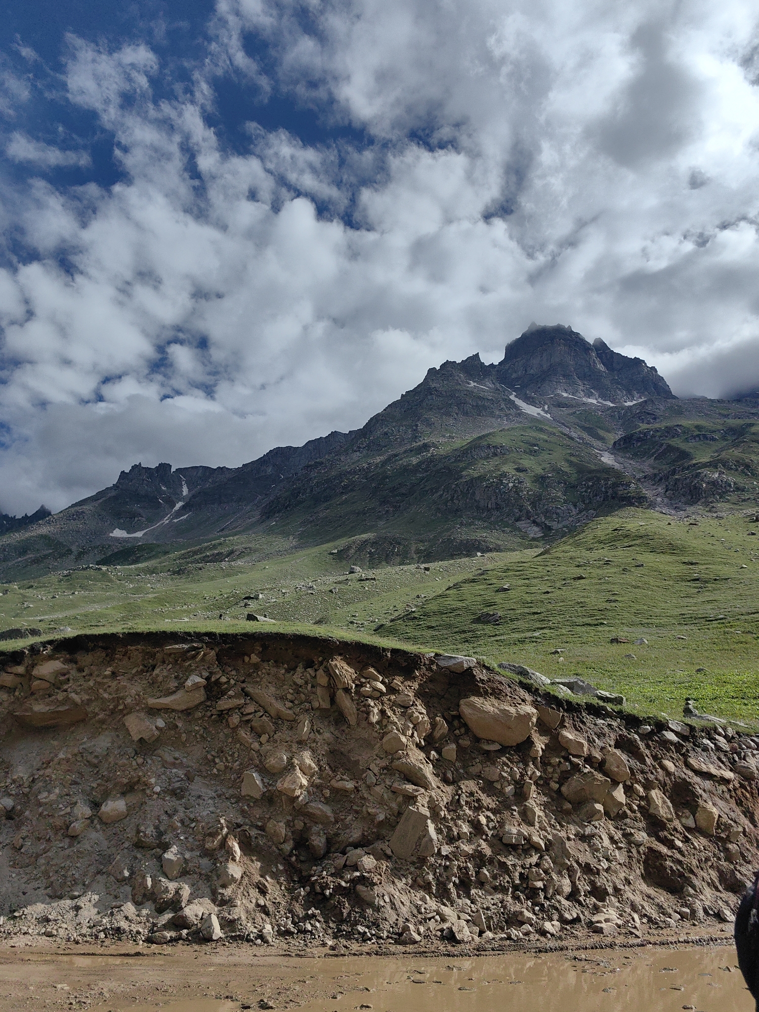 A vast, mountainous landscape unfolds under a partly cloudy sky. In the foreground, a muddy track with puddles lies at the bottom of the frame. To the left, a steep, eroded embankment reveals layers of soil and scattered rocks, topped with a strip of green grass. Behind this, rolling green hills rise, dotted with rocks and patches of snow. The background is dominated by a rugged, dark mountain peak shrouded in mist at its summit. The scene is devoid of people or any discernible structures. The overall impression is one of remote, natural grandeur. The location is identified as the city of Bashisht, India.