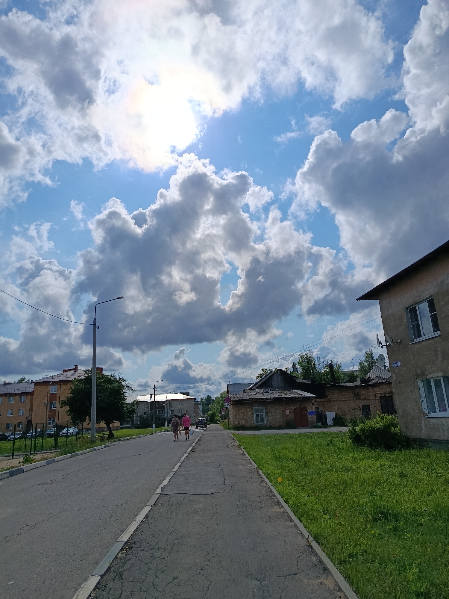 The image captures a sunlit street scene in Vareya, Russia, dominated by a dramatic sky filled with voluminous, billowing white and gray clouds, with the sun peeking through the upper center. A paved road with visible cracks and a concrete curb stretches into the distance, bordered by a well-maintained grassy verge on the right. On the left side of the road, a sidewalk runs parallel to the street, with two individuals walking away from the camera. Further down the road, several buildings are visible, including apartment buildings and older brick structures with pitched roofs. A single lamppost stands prominently near the left sidewalk. Cars are parked on the far left side of the street, in front of an apartment complex. The environment appears to be a typical urban or suburban setting in a Russian town.