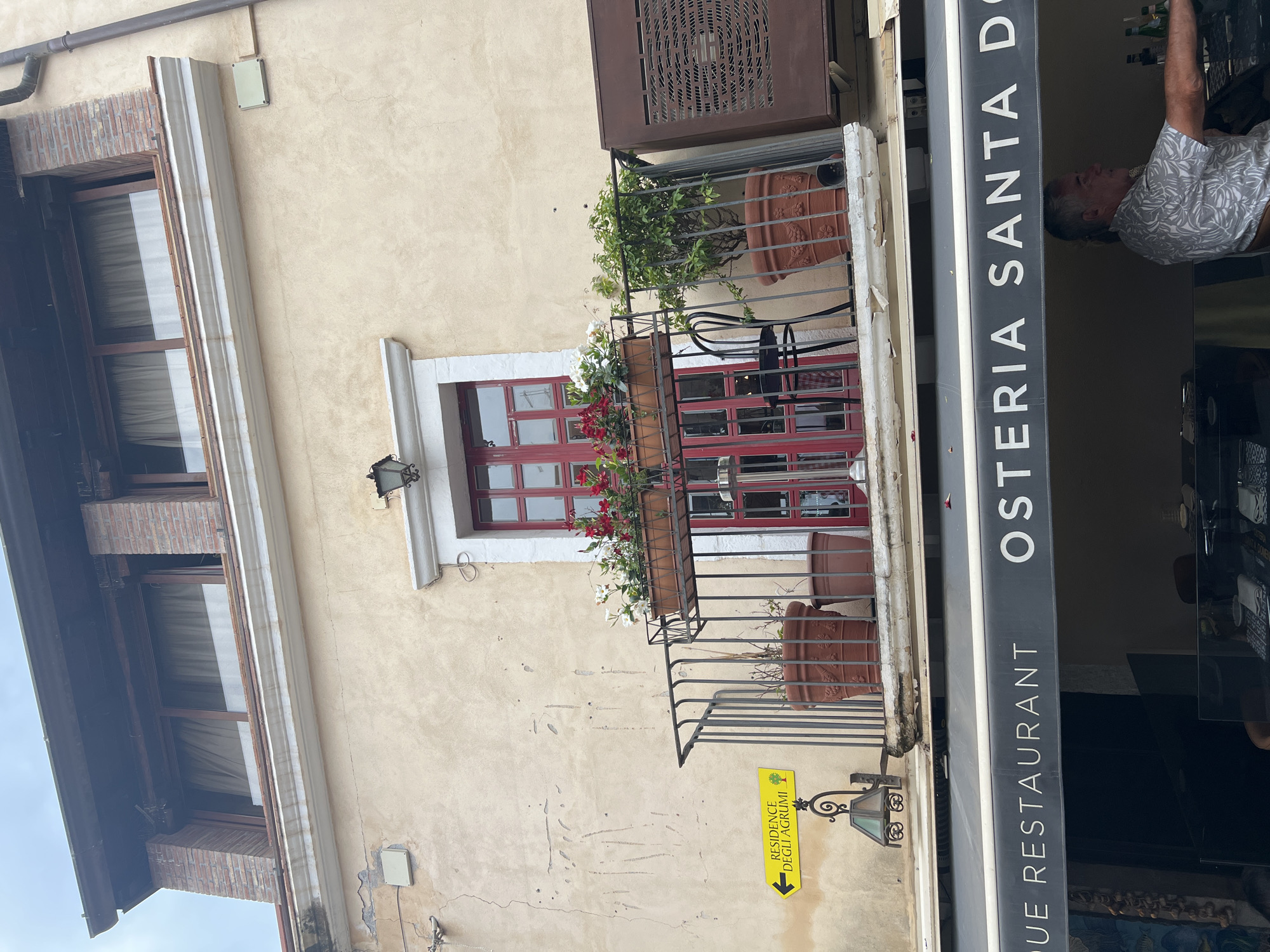 The image shows the exterior of a restaurant in Taormina, Italy, named "Osteria Santa Do". The building facade is beige and weathered, with a balcony on the second floor featuring a red-paned window, a black metal railing, and several potted plants. A yellow directional sign pointing left reads "RESIDENCE DEGLI AGRUMI" with a small tree icon. Below the balcony, a dark awning displays the restaurant's name in white lettering. A man in a floral shirt can be seen from the chest up in the lower right portion of the frame, behind a counter with bottles. The sky is overcast.