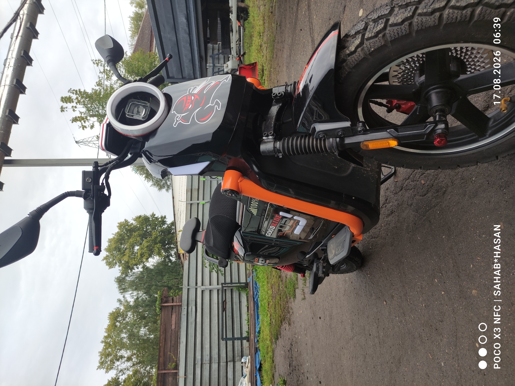 A black and orange electric scooter is parked on a gravel surface outdoors, with a chain-link fence and wooden structures in the background. The scooter has a circular headlight with a white rim and a red inner ring. A red and white graphic of a masked figure wielding swords is displayed on the front panel. The scooter features knobby tires, red brake calipers, and a black seat with an orange frame visible. The ground is a mixture of dirt and small stones, with some patches of green grass visible behind the fence. The sky is overcast and cloudy. A timestamp in the lower right corner reads "17.08.2026 06:39", and a watermark in the lower left corner indicates "POCO X3 NFC | SAHAB*HASAN". There are no people visible in the image. The environment appears to be an outdoor storage area or a yard.