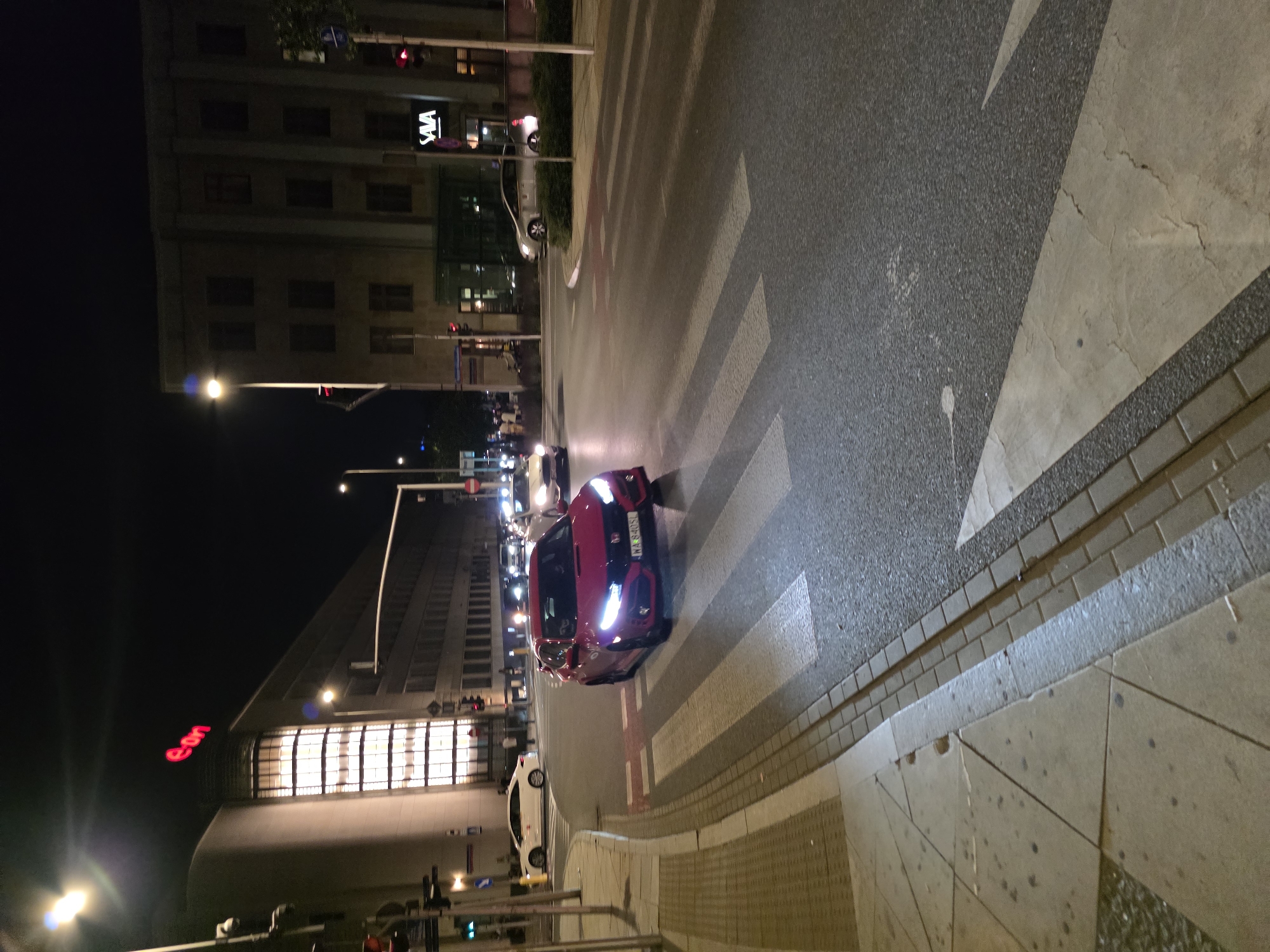 The image shows a street scene at night in Warsaw, Poland, likely near the Eon building. A red Honda Civic is driving over a pedestrian crossing. The car has its headlights on and appears to be the main subject of the image. In the background, there are several buildings, one of which has the "eon" logo illuminated in red. Another building has the "SAVA" logo visible. Traffic lights and street lamps are also present, indicating an urban environment. The road is wet, suggesting recent rain or damp conditions.