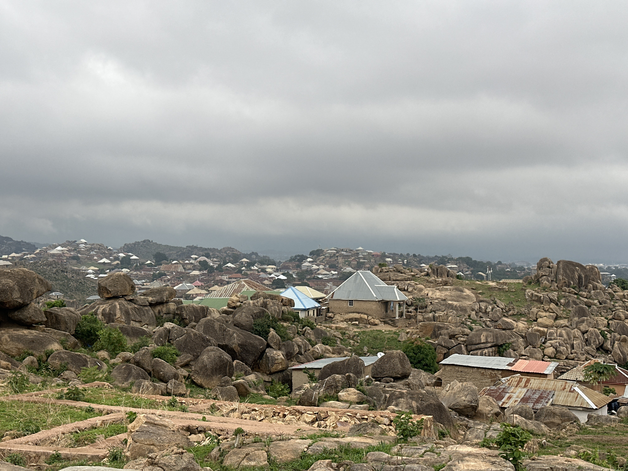 The image shows a landscape of Jos, Nigeria, characterized by rocky terrain interspersed with residential buildings. The foreground features numerous large, rounded boulders with patches of grass and low vegetation. Several buildings with corrugated metal roofs are visible among the rocks, ranging in color from silver to orange and green. In the mid-ground, more houses are situated on a hillside, extending into the distance under an overcast sky. The sky is uniformly gray, suggesting a cloudy or rainy day. The architecture appears simple and functional, blending into the natural rocky environment.