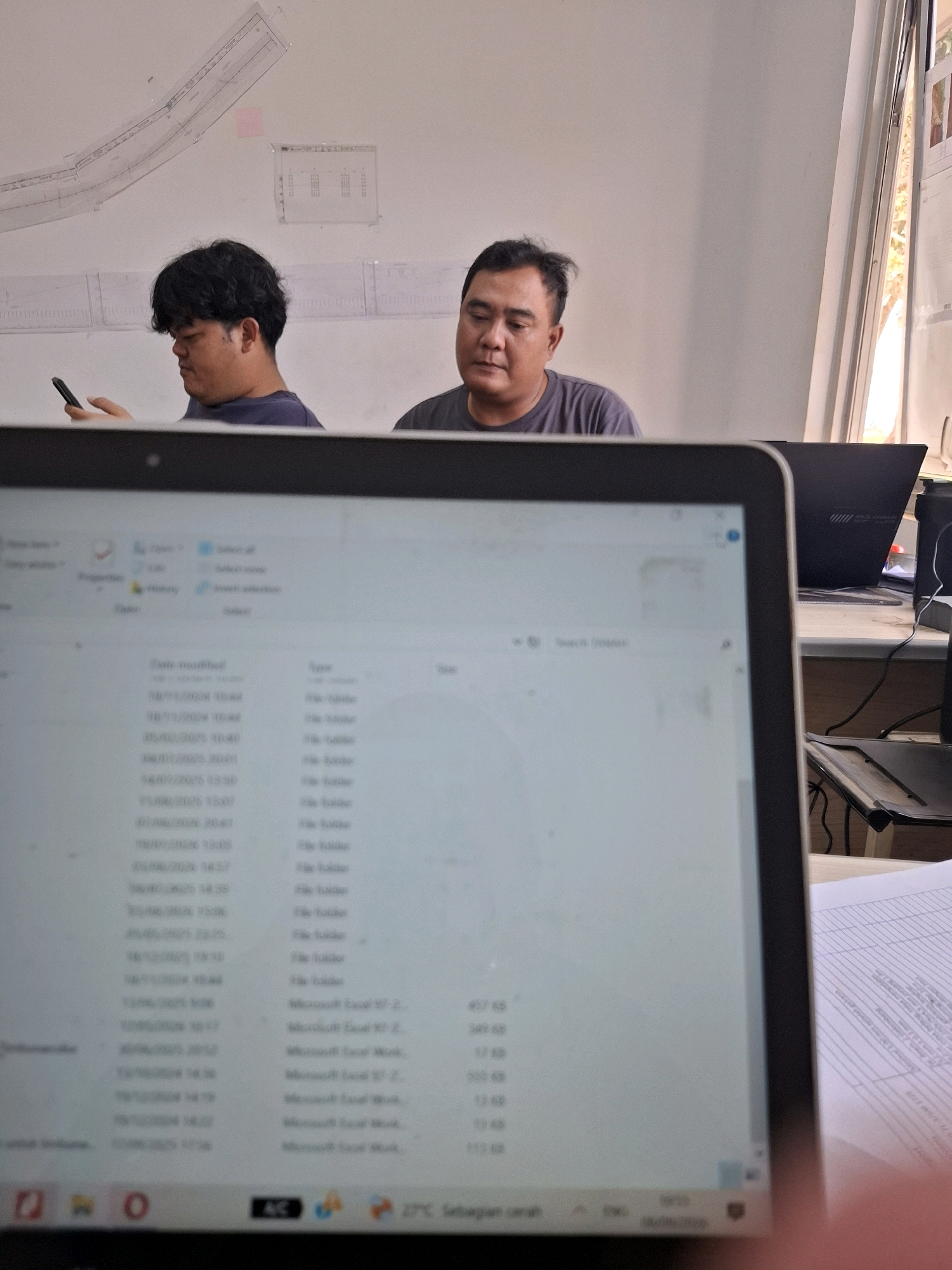 The image shows two men sitting at desks in what appears to be an office or workspace. The man on the left is looking down at his phone, while the man on the right is looking forward. In the foreground, a laptop screen displays a file directory with a list of files and folders. On the wall behind the men, there are some technical drawings or blueprints. The environment appears to be well-lit, with natural light coming from a window on the right side of the frame. The overall impression is one of a casual work environment. Based on the context provided, this scene is located in the city of Ciheulang Tonggoh, Indonesia.