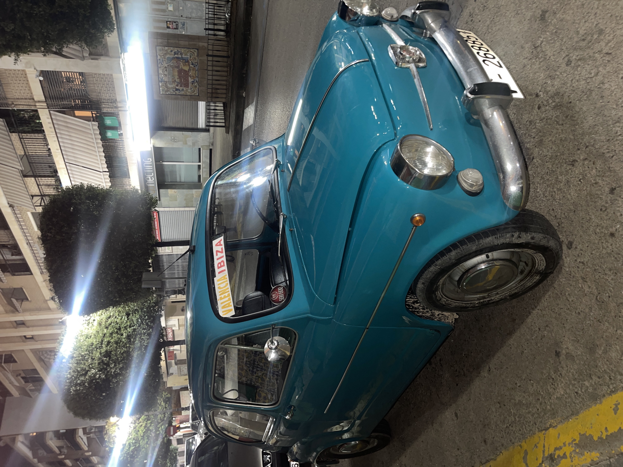 This nighttime image captures a turquoise vintage Seat 600 car parked on Carrer de la Sort, Manises, Spain, specifically in front of the building housing the "Necting" office (Carrer de la Sort, 40). The vehicle is positioned on the right side of the street, with its front facing toward the viewer's right.
The car, a classic Seat 600, is a bright turquoise or teal color, featuring chrome accents including bumpers, headlight surrounds, and side trim strips. A sticker displaying "60 SEAT 600" in a red circle is visible on the dashboard, and a yellow banner with "VALENCIA IBIZA" in red letters is affixed to the inside of the windshield. The front license plate displays "268851". The car's tires and lower body panel exhibit splashes of dirt or mud.
The scene is illuminated by strong artificial streetlights, casting bright lens flares across the upper left and center of the image and creating reflections on the car's paintwork. The street surface is dark grey asphalt, with a faded yellow painted line visible along the curb on the left side.
In the background, multi-story buildings line the street, typical of Spanish urban architecture. These buildings feature balconies with black railings and light-colored, striped retractable awnings. On the ground floor, a business named "Necting" is visible with a modern, white-fronted facade and glass windows. Adjacent to "Necting" is another closed storefront with a corrugated metal roll-down security door. To the right of the "Necting" store, a decorative, colorful tiled mural is affixed to a building wall, partially obscured by a black metal fence. Neatly trimmed, spherical green trees line the street, partially illuminated by the streetlights. Another vehicle, a silver or grey car, is partially visible parked behind the turquoise Seat 600 on the left. No individuals are present in the image.