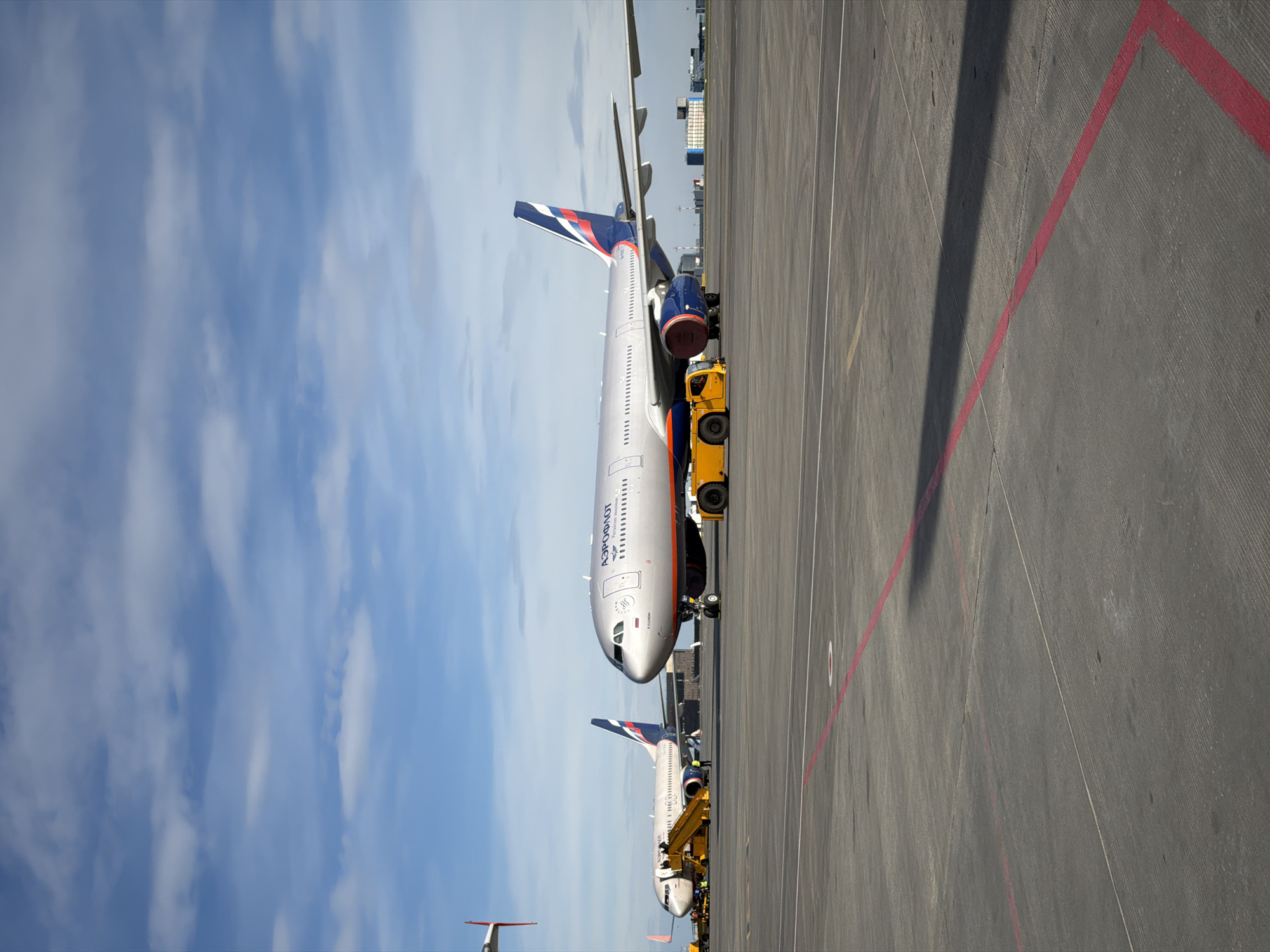 A wide, correctly oriented shot captures two Aeroflot passenger airplanes parked on an airport tarmac under a bright, partly cloudy blue sky. The scene depicts routine daytime airport operations in Khimki, Russia.
In the foreground and mid-ground, a large, light grey passenger aircraft, identifiable by the Cyrillic "АЭРОФЛОТ" (Aeroflot) lettering on its fuselage, is prominent. A small Russian flag is painted near the cockpit windows. A bright yellow pushback tug is attached to the nose landing gear of this aircraft, indicating it is either being pushed back from a gate or towed across the apron. The aircraft's tail fin displays the distinctive blue, red, and white Aeroflot livery. One of its large engines, also featuring blue and red accents, is visible near the tug.
Further in the background and to the right, a second, smaller Aeroflot aircraft is partially visible. It also appears to be accompanied by yellow ground handling equipment, suggesting similar operational activity.
The airport tarmac is a grey concrete surface, marked with distinct red and white lines that delineate operational zones. Shadows from the aircraft and possibly other airport structures stretch across the ground. In the distance, under the clear sky, some low-rise buildings and structures, typical of airport infrastructure, are faintly visible.