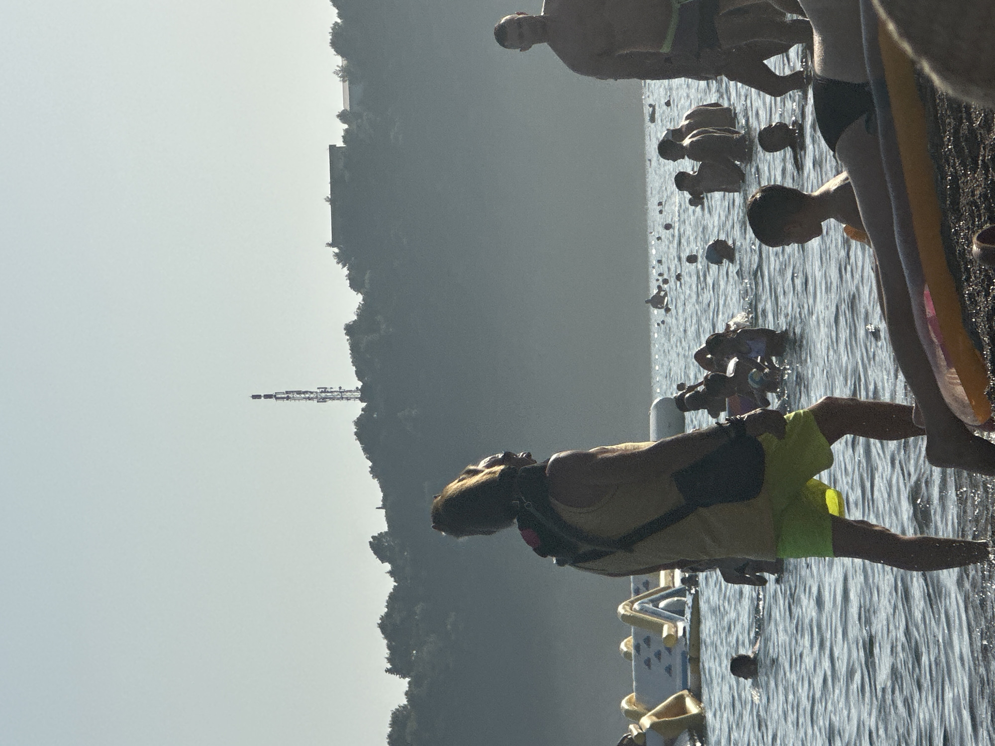 The image captures a crowded beach scene in Boreti, Montenegro. The water is populated with numerous individuals swimming, wading, and playing. In the foreground, a person wearing a yellow tank top and neon green shorts stands facing away from the camera, with a black bag slung over their shoulder. Another person, partially visible in the lower right corner, is lying on a float. In the mid-ground, a group of people are scattered throughout the water, with some appearing to be near inflatable water toys. The background features a densely wooded hillside topped with a building and a telecommunications tower. The sky is hazy, suggesting a bright, sunny day.