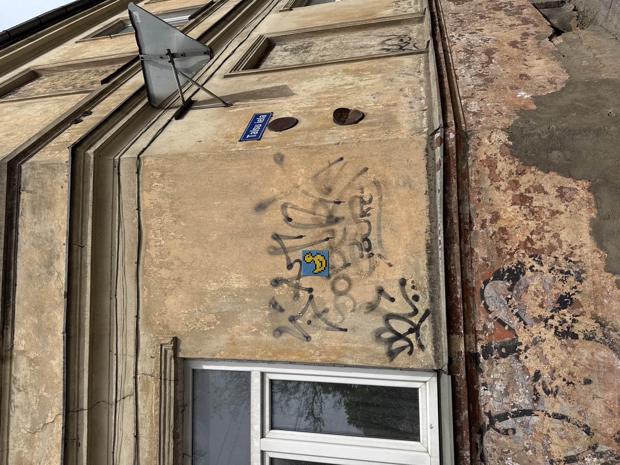 The image shows the exterior of a building in Riga, Latvia. A satellite dish is mounted high on the wall, above a street sign indicating "Taksu iela". Below the sign, there are two dark, circular metal objects affixed to the wall. Spray-painted graffiti surrounds a blue and yellow pixel art figure resembling a Pac-Man ghost. A white-framed window is visible below the graffiti. The building's façade exhibits signs of age and disrepair, with peeling paint and visible cracks. The adjacent section of the building on the right has extensive paint loss, exposing the underlying layers. Electrical wires run vertically along the left side of the frame.