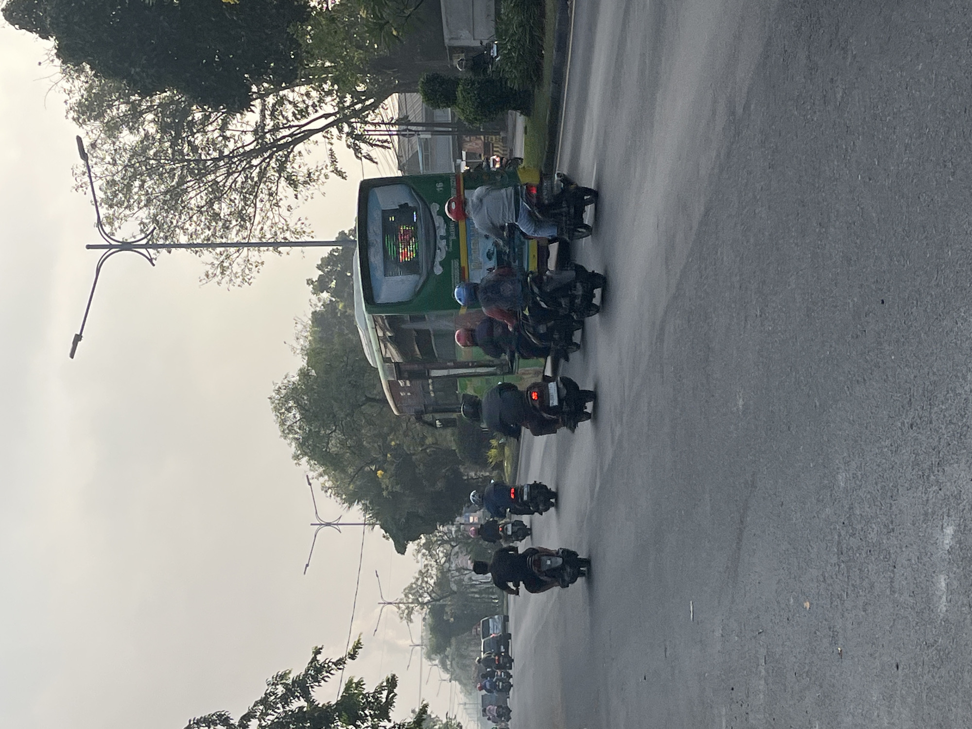 The image captures a street scene in Gelam, Indonesia, with a prominent green bus driving on the right side of the road. Several motorcycles are also on the road, with riders in various positions. The sky is overcast and hazy, suggesting a diffused light or possibly pollution. Trees line the sides of the road, and streetlights are visible, indicating an urban or semi-urban environment. The road surface is asphalt, and there appear to be some faint tire marks. The overall impression is of a typical, busy day on a road in Indonesia.