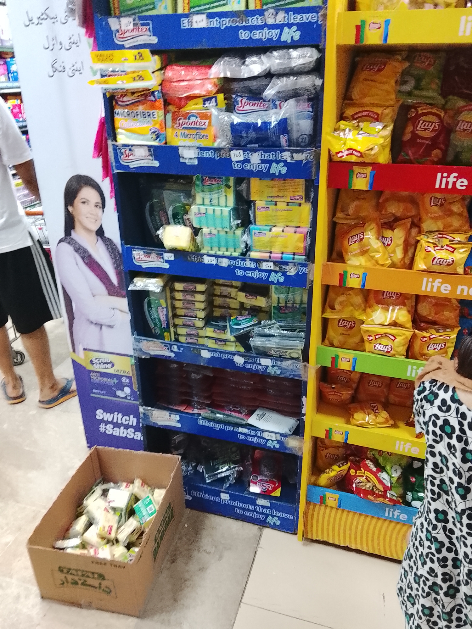 The image captures a scene inside a retail establishment, likely a grocery store or supermarket, located in Gulgasht Colony, Pakistan. Two people are partially visible: a man in casual attire with bare legs and slides, and a person in a black and white patterned dress. The environment is characterized by organized displays of merchandise.
A blue shelving unit is stocked with cleaning products, predominantly Spontex brand items such as microfiber cloths and scrub pads. The shelving unit is decorated with the slogan "Efficient products that leave to enjoy life". A promotional standee featuring an image of a woman accompanies the display, along with a slogan that includes the hashtag #SabSa.
Adjacent to the blue shelving unit is a yellow shelving unit filled with Lay's potato chips. The displays are labeled with the word "life." A box containing individual bars of soap is located on the floor in front of the shelving units, marked with text in Urdu and the English words "FREE TRAY".
