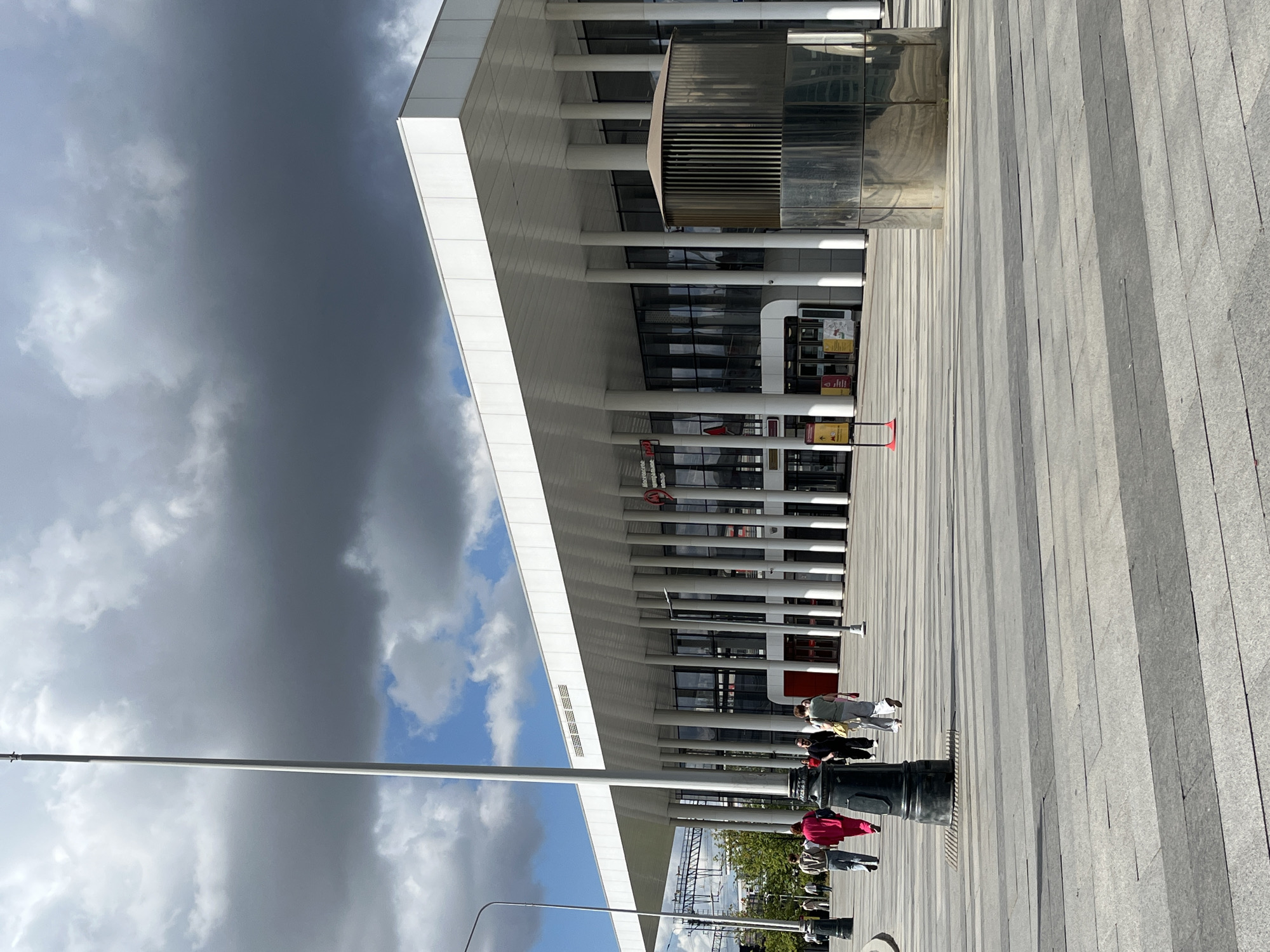 The image captures the exterior of the Moskovsky railway station in Moscow, Russia. The building features a modern architectural design with a white, overhanging roof supported by numerous vertical white columns. Large glass windows line the facade, reflecting the sky and clouds. In front of the station, a paved plaza is visible, with people walking across it. A prominent dark lamppost stands on the left side of the image, and a cylindrical metallic structure with a slatted top is located on the right. The sky is partly cloudy, with dramatic dark clouds dominating the upper portion of the frame, contrasted by patches of blue sky.