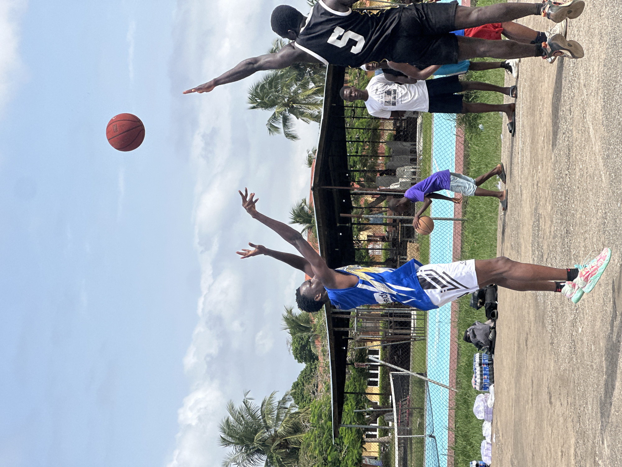 A medium shot captures a basketball game in progress on an outdoor court. The sky is partly cloudy, with palm trees and a building visible in the background. In the foreground, two young men are actively playing, one in a blue and white jersey, the other in a black jersey with the number 5. The man in the blue jersey is in mid-air, reaching for the ball, which is also in the air. The man in the black jersey is also jumping, attempting to block. Other individuals are observing the game from the sidelines. The court appears to be made of concrete, with a net and pool in the background. Bottles of water and other items are arranged near the edge of the court. The overall scene suggests a casual basketball game in a tropical or subtropical environment.
