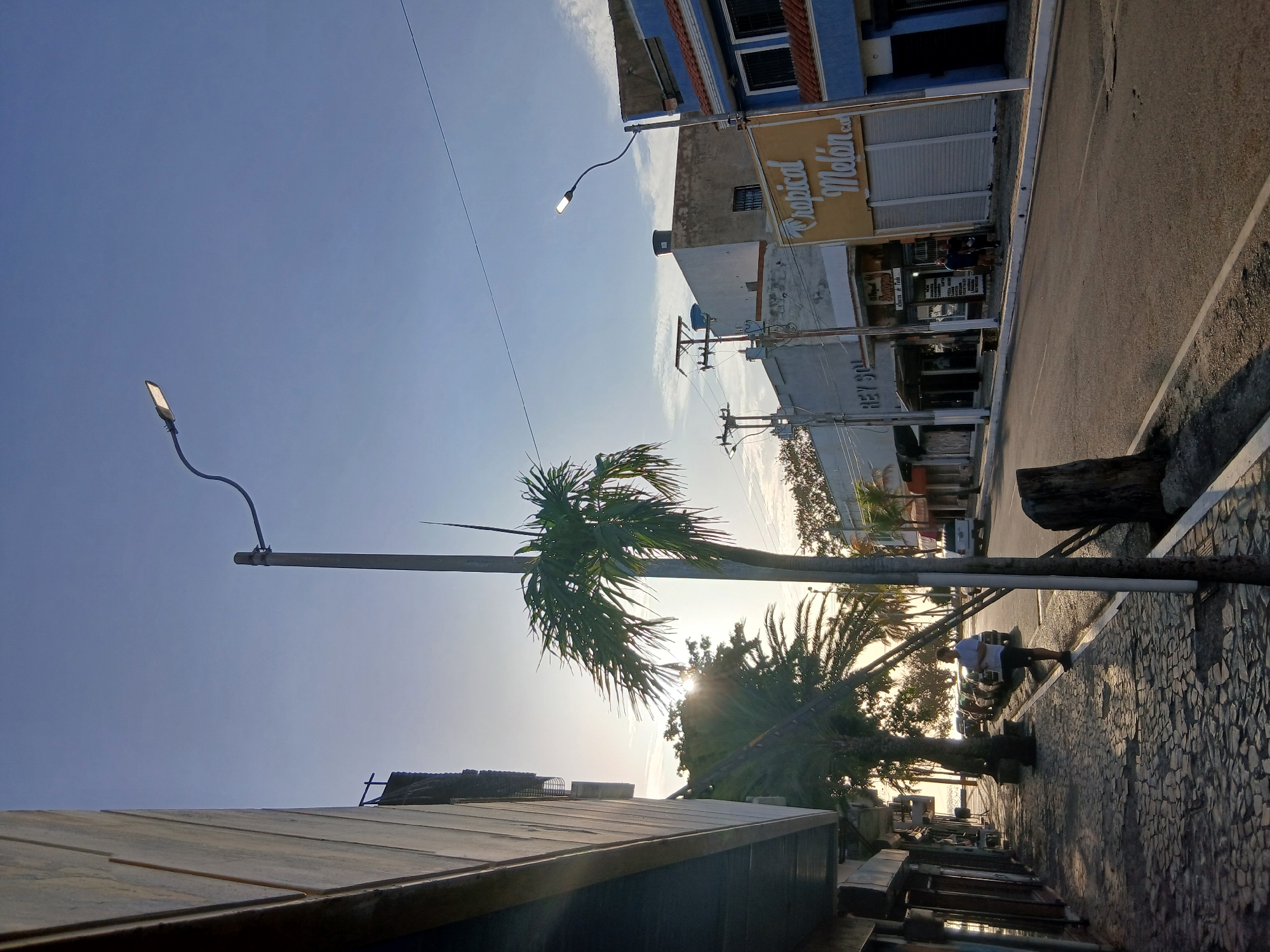 A view from a street in Juan Griego, Venezuela, angled to the right. The sky is blue. Buildings, most of which are closed, line one side of the street. On one building, a sign reads "Tropical Melon." A palm tree and street light stand between buildings. A man in light clothing and dark shorts leans against a palm tree on the street. Cars are parked along the road.