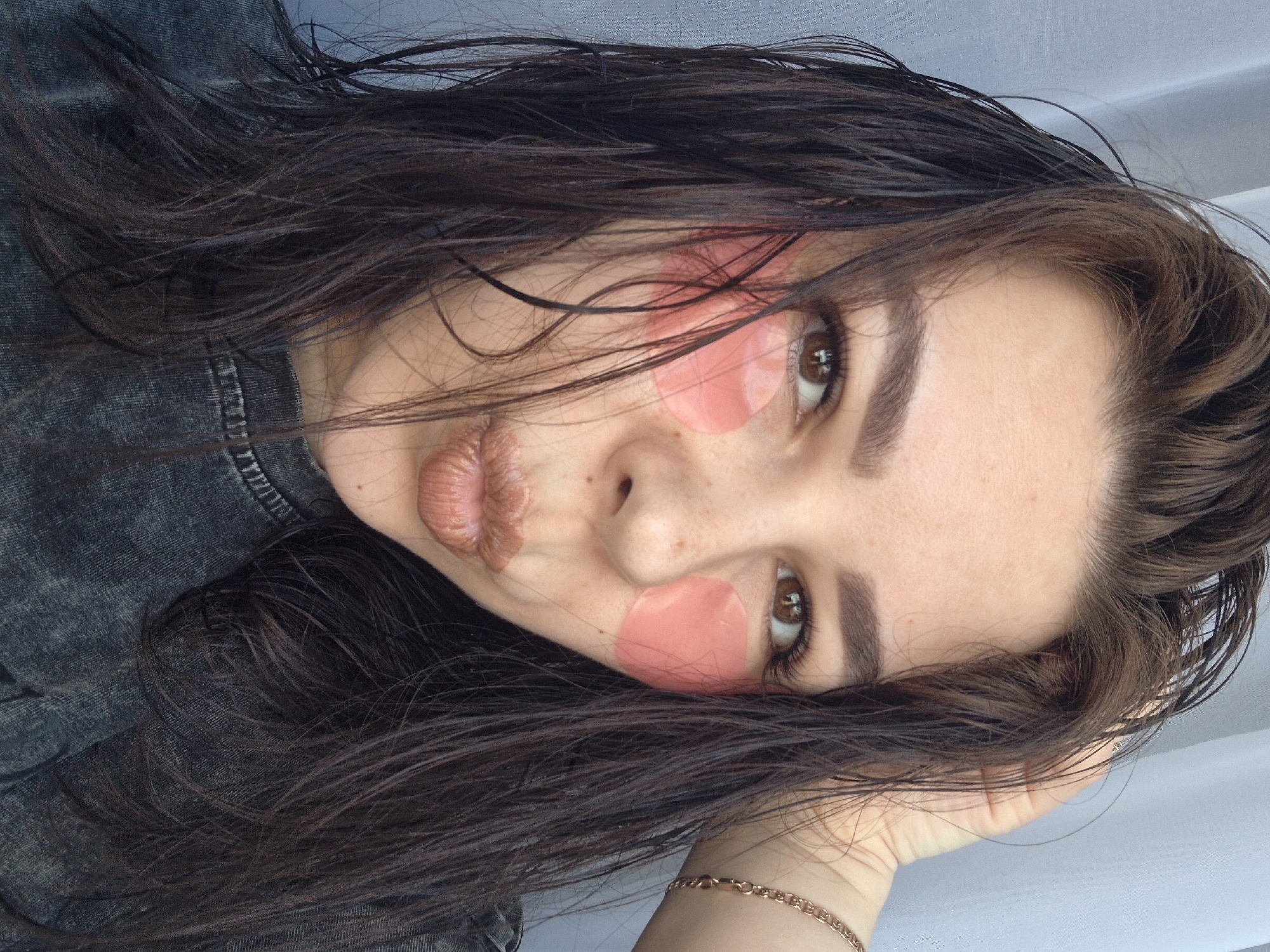 The image presents a close-up, angled portrait of a young woman, rotated 90 degrees as if she is lying down or resting her head, potentially in Tobolsk, Russia. Her long, dark, slightly wet-looking hair frames her face, with strands falling across her forehead and cheeks. She looks directly at the viewer with intense brown eyes, her well-groomed eyebrows prominent.
A distinctive feature is the presence of two oval-shaped, translucent pink patches on her face: one positioned under her left eye and another on her right cheek/temple area, indicative of a skincare mask or a decorative element. Her lips are full and sculpted into a subtle pout, highlighted by a light, nude-toned lipstick. She appears to be wearing a dark, textured garment, possibly denim, which is visible on her shoulder and part of her arm. Her right hand, adorned with a delicate gold chain bracelet, supports her head in the lower right corner of the frame. The background is a plain, light-colored surface, suggesting an indoor setting with soft, ambient lighting. No text is visible. The overall impression is one of relaxed intimacy or a moment captured during a beauty routine.