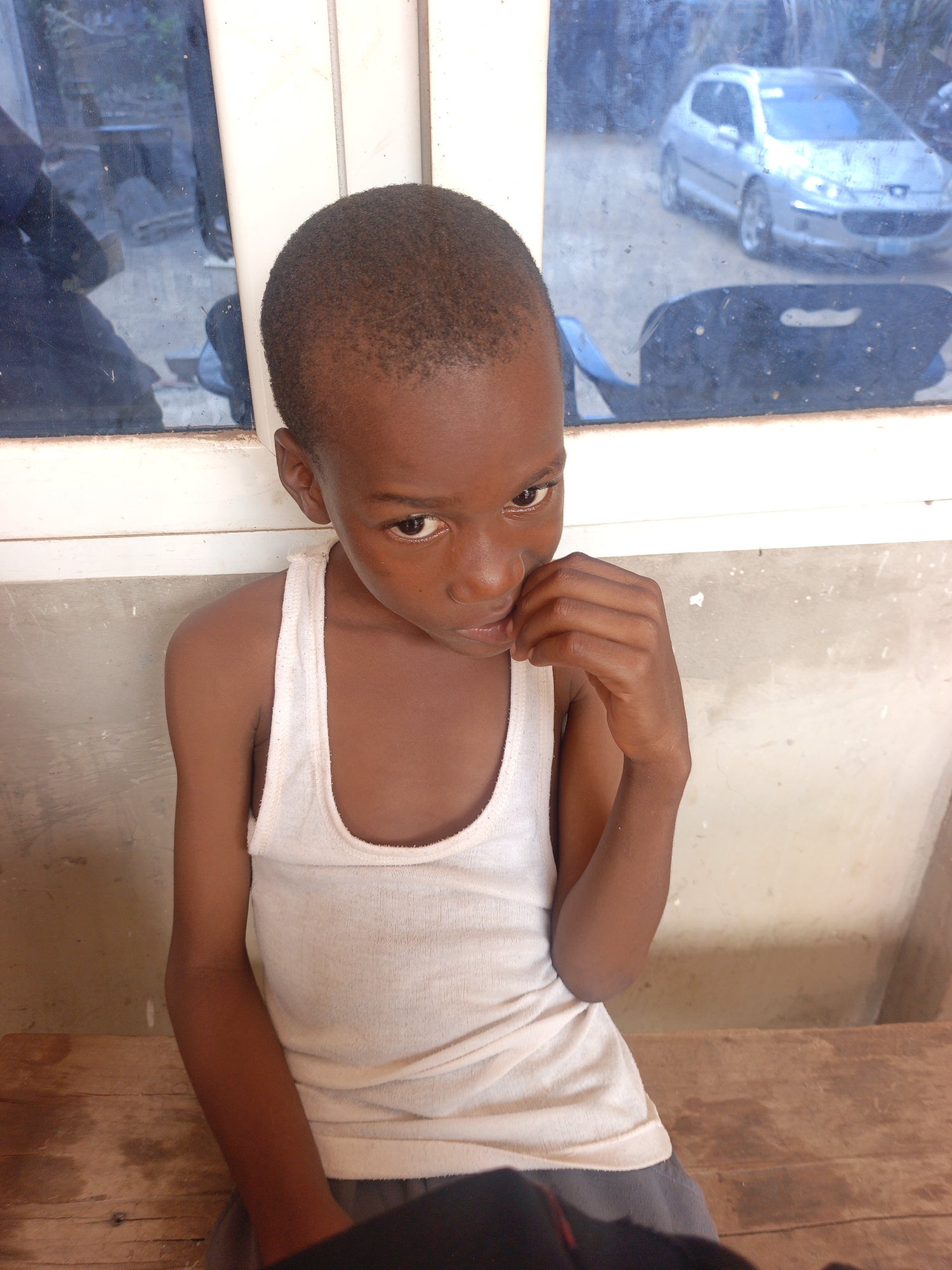 A young, dark-skinned boy is seated on a wooden bench inside a room with a cement wall. He is wearing a white tank top and dark-colored shorts. He is looking directly at the camera with his right hand near his mouth. Behind him is a window with two panes; the reflection in the window shows a silver car and other blurred objects. The location is presumed to be Akwanga, Nigeria.