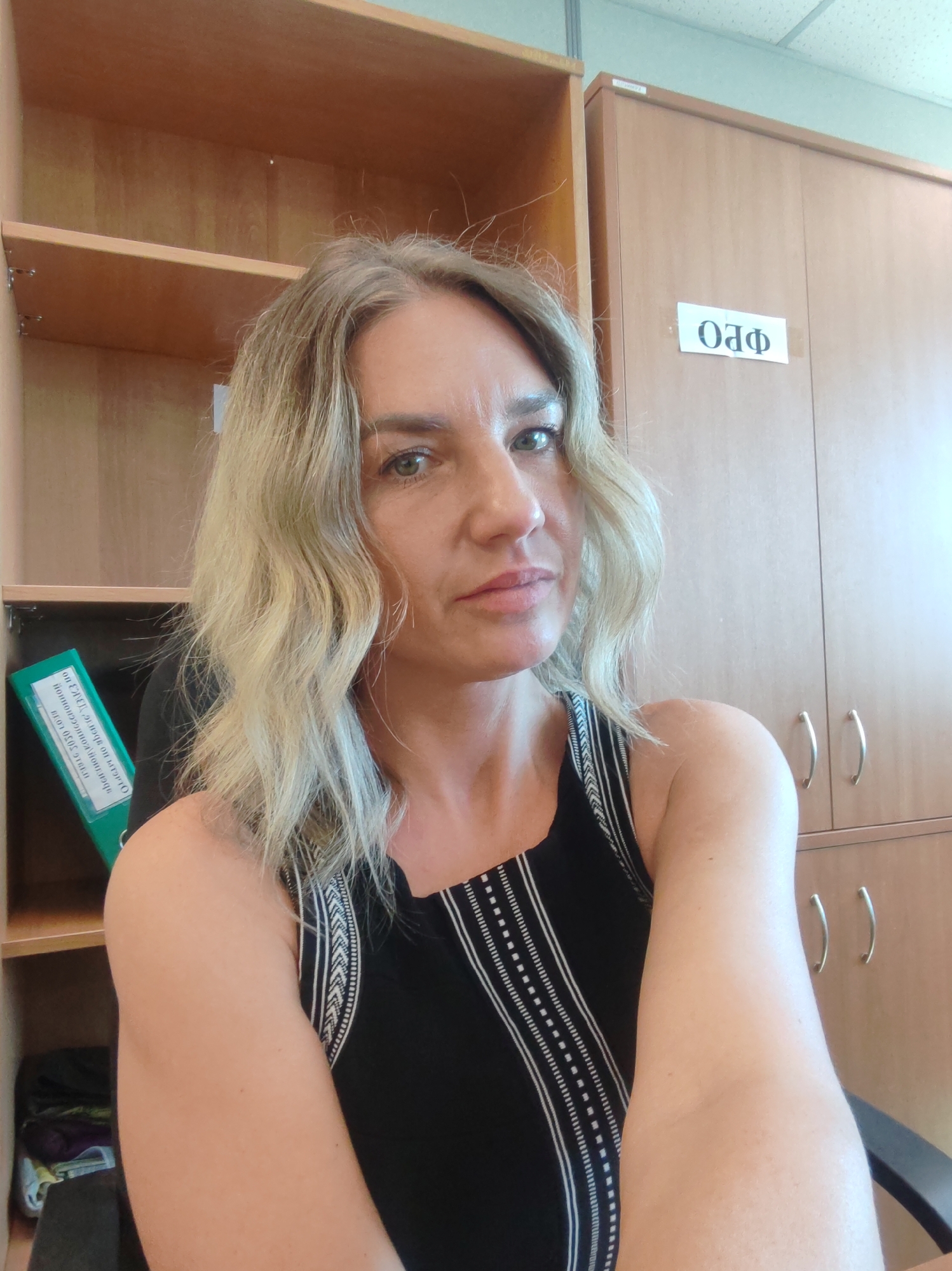 A woman with light blonde wavy hair is in a room with light brown wooden cabinetry. The woman has green eyes and is wearing a sleeveless black top with white striped detailing. Behind her on the left is shelving with a green binder. On the right is a closed cabinet with silver handles and a white sign with Russian text. The location is likely Petropavlovsk-Kamchatsky, Russia based on the presence of Russian text.