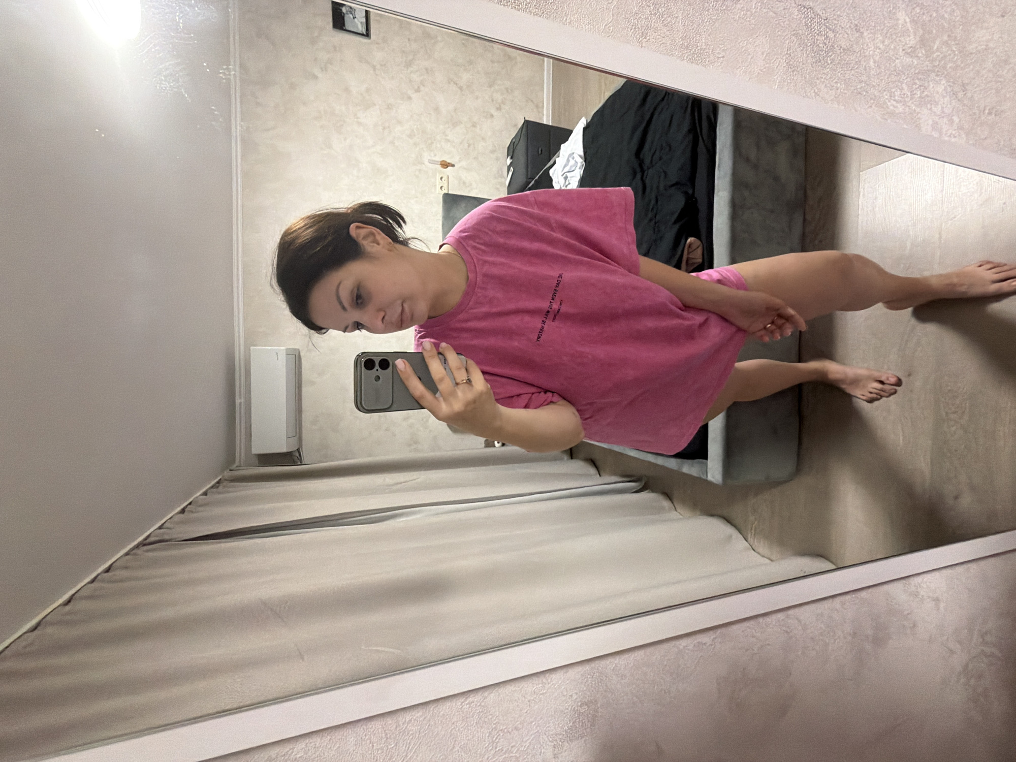 A woman stands in front of a mirror in a room, taking a selfie. She is wearing a pink t-shirt and has her hair pulled back. The room appears to be a bedroom, with a bed and curtains visible. There is also an air conditioner mounted on the wall. The image is taken from a low angle, showing the woman from the knees up. The location is identified as Sochi, Russia.
